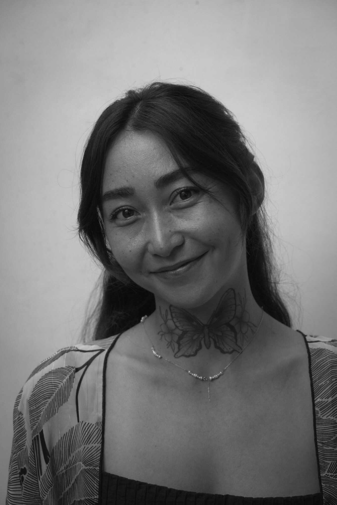Woman with long dark hair smiling, wearing a patterned top and a necklace, against a plain background.
