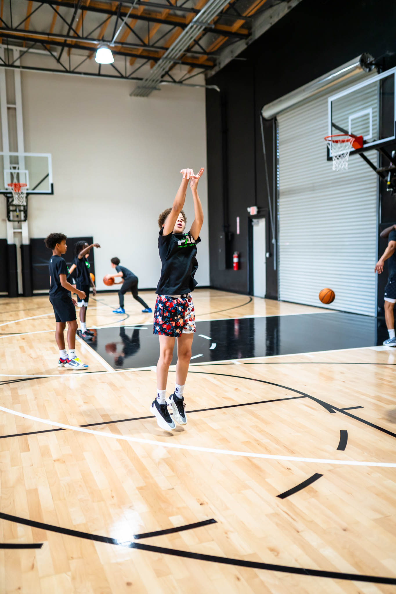 Children playing basketball in an indoor gym, one girl shooting the ball, others watching and playing.