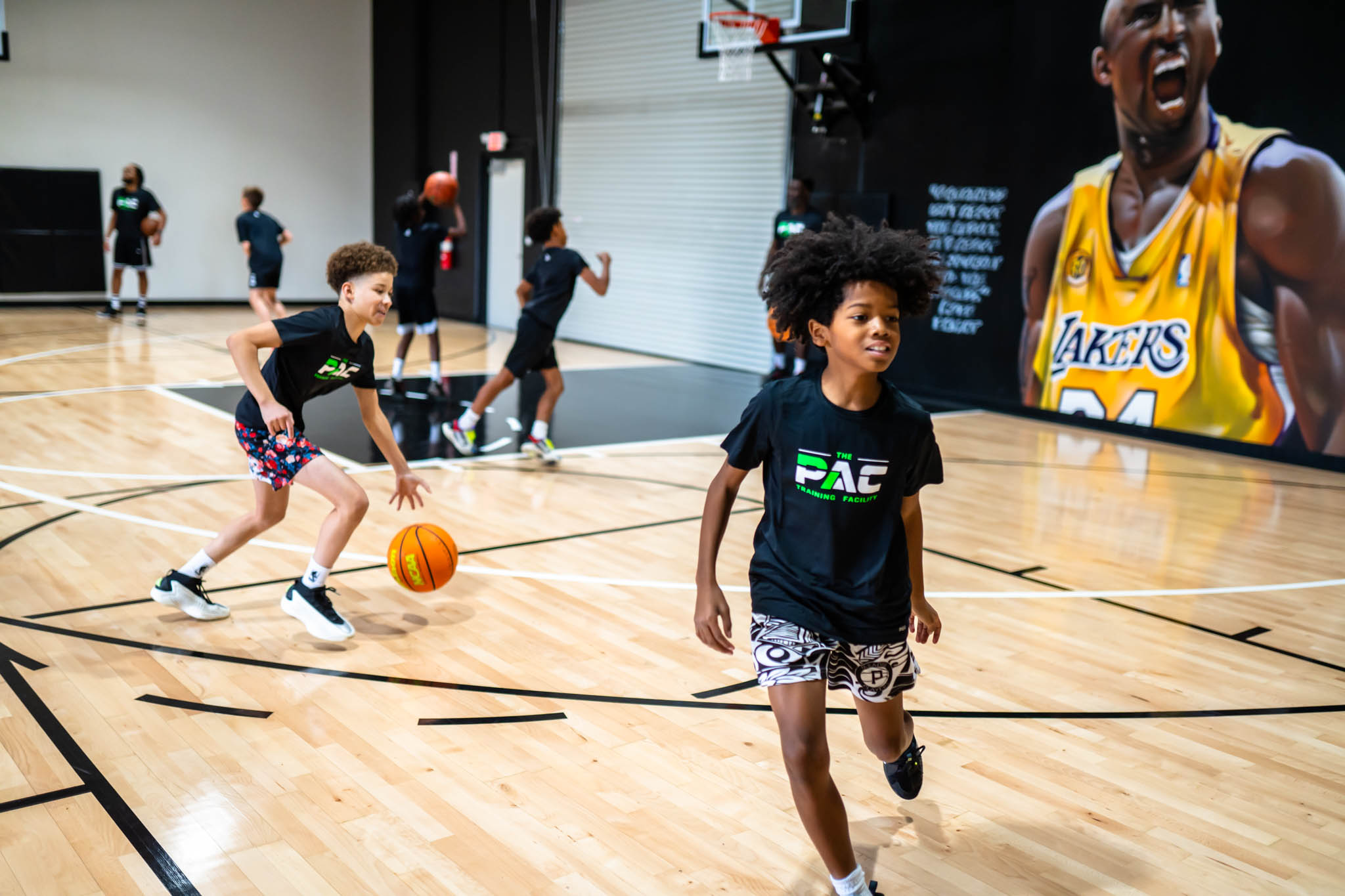 Children playing basketball on an indoor court with a large screen showing a basketball player, some children dribbling, smiling, and running.