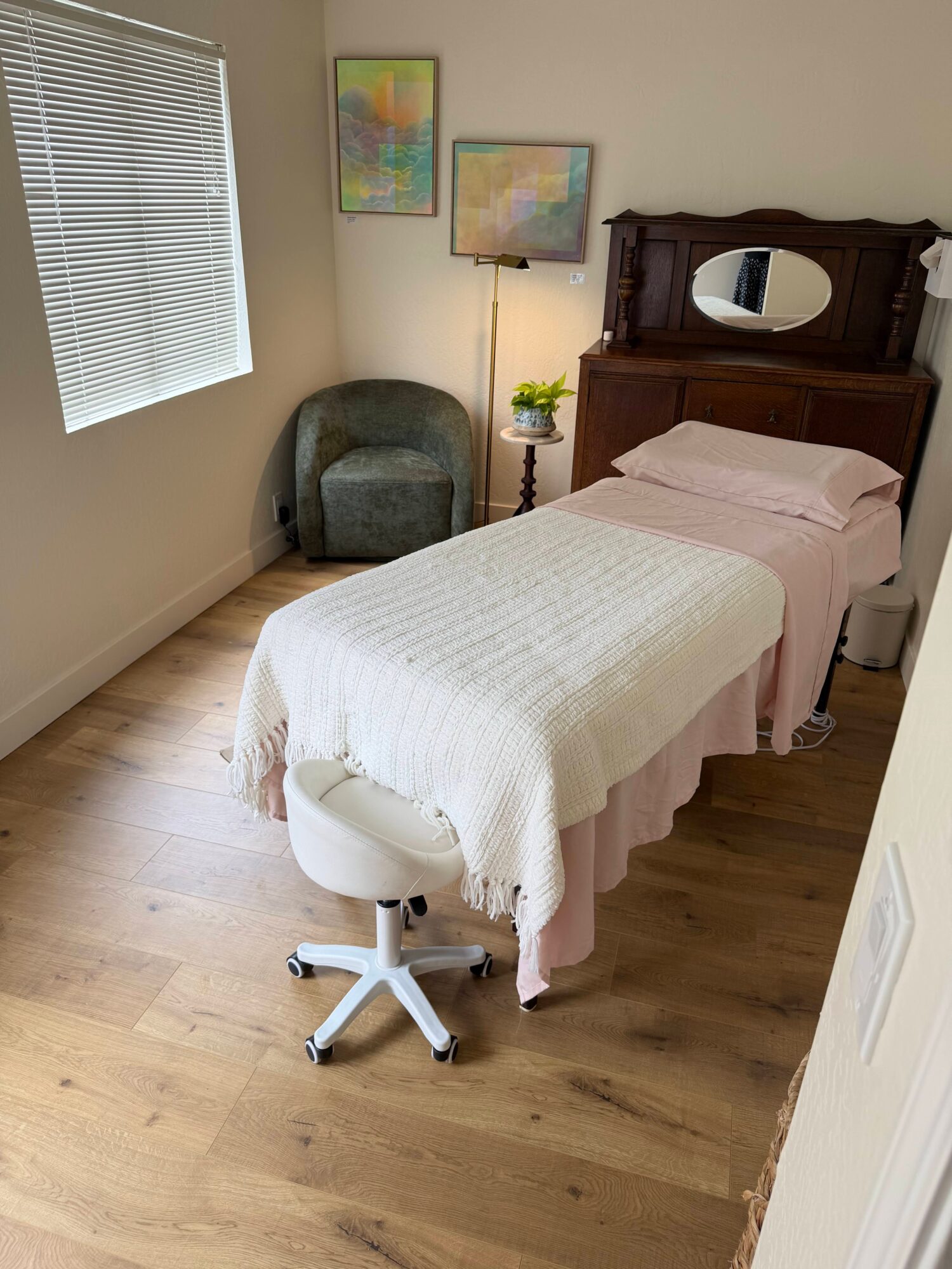 Room with bed, chair, window with blinds, and wall art, featuring wooden floor and a mirror on the headboard.