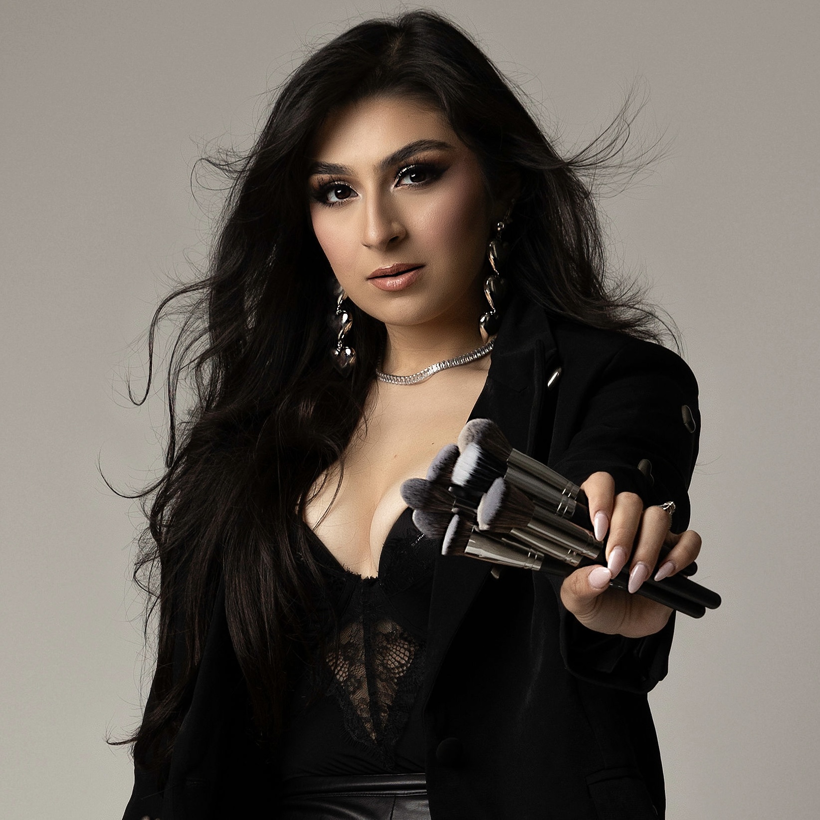 Woman with long dark hair holding a gun, wearing black lace top and blazer, against a plain background.