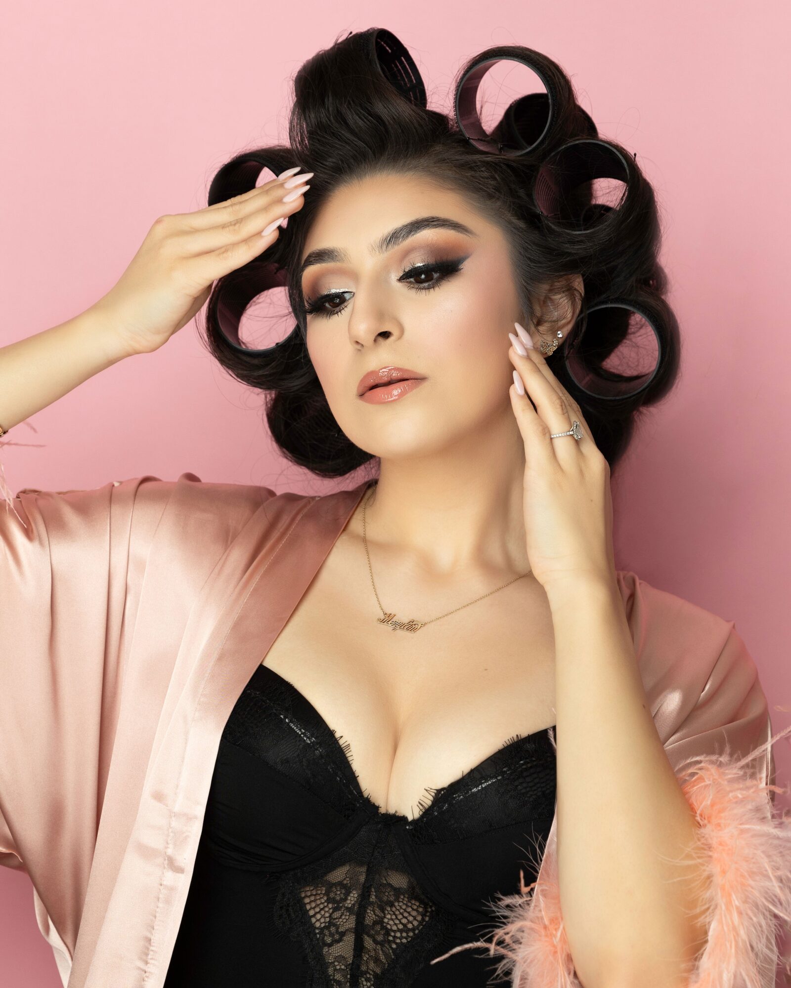 Woman with styled hair, makeup, and jewelry poses against pink background, wearing a black top and satin robe.