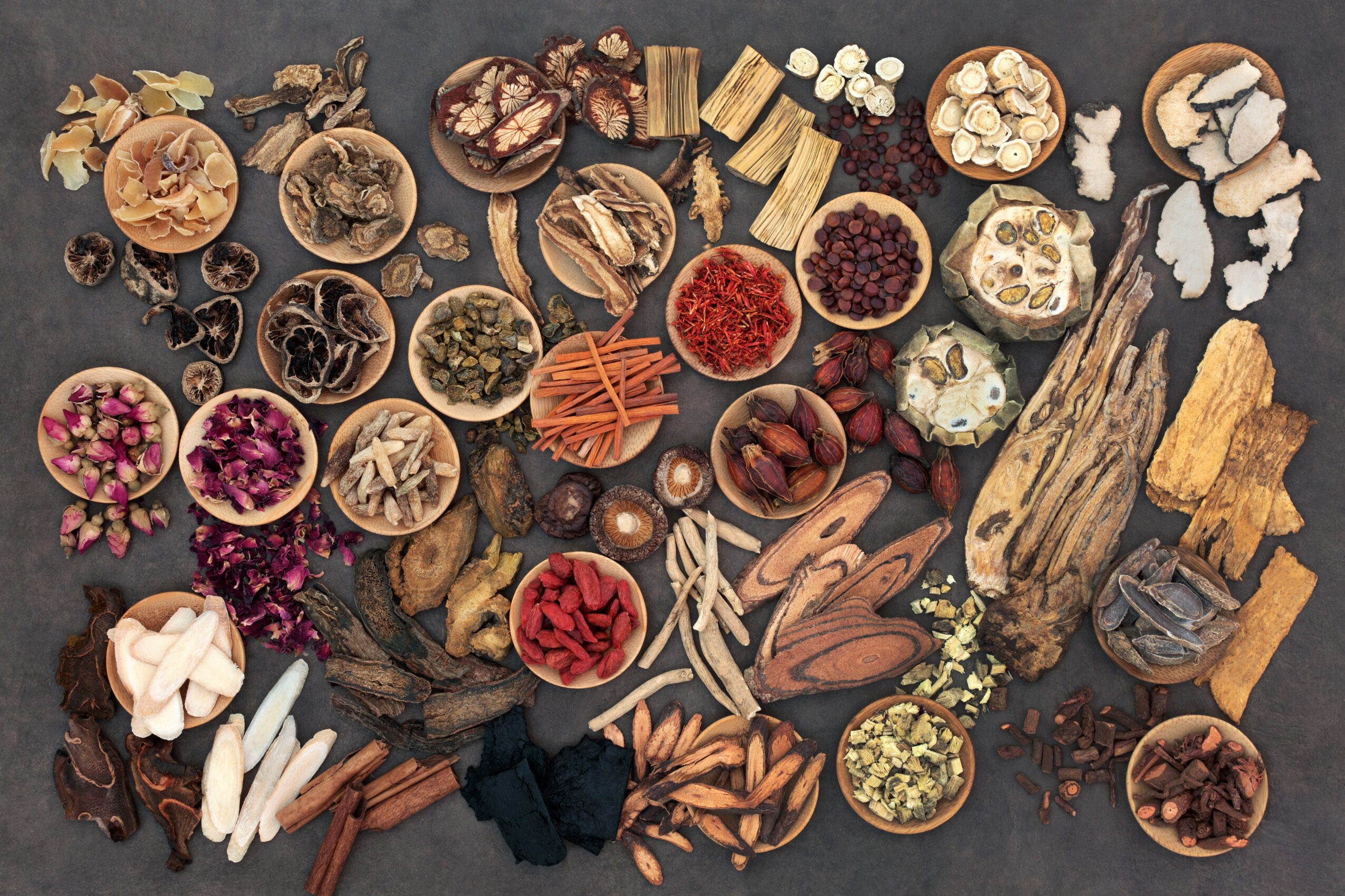 Assorted herbs, spices, and dried botanicals arranged on a dark surface with wood pieces and small bowls.