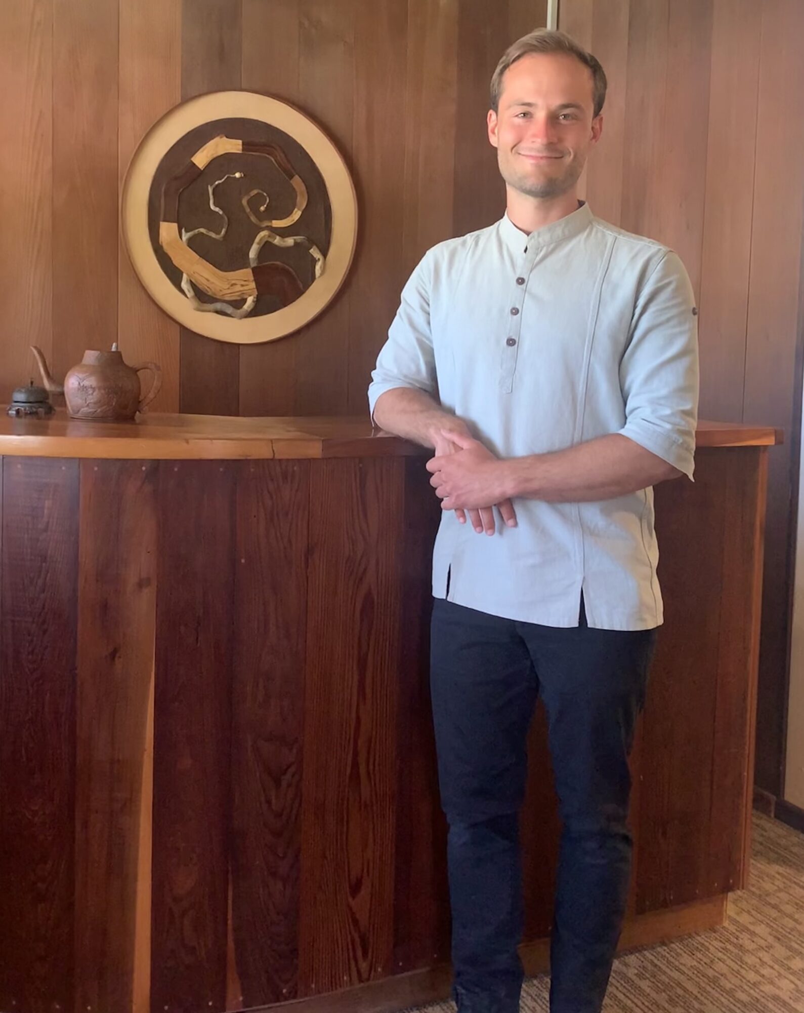 Man standing indoors with wooden wall and artwork, smiling, wearing a light blue shirt and dark pants.