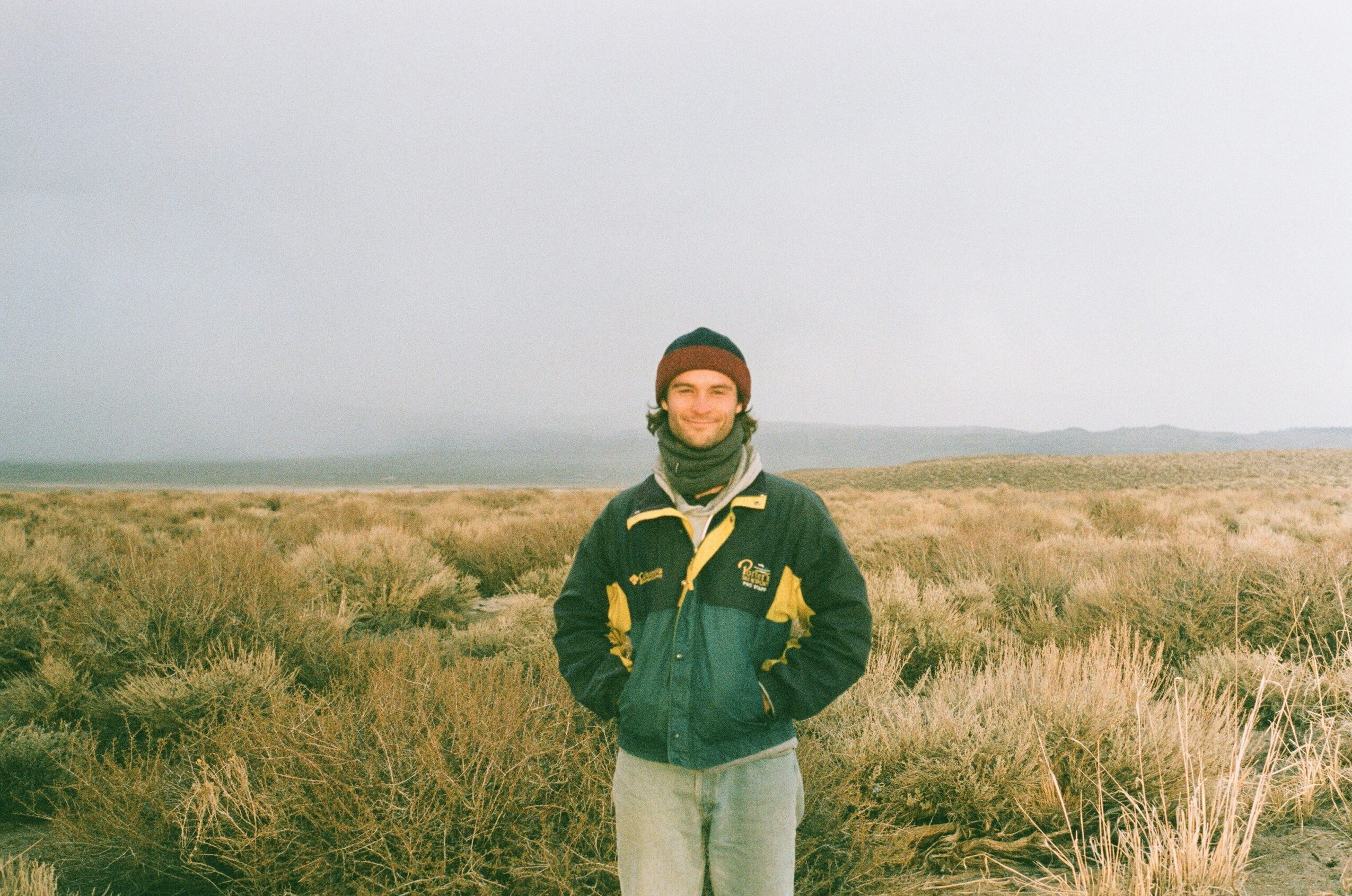 Person standing outdoors in a grassy field under a cloudy sky, wearing a jacket and beanie.