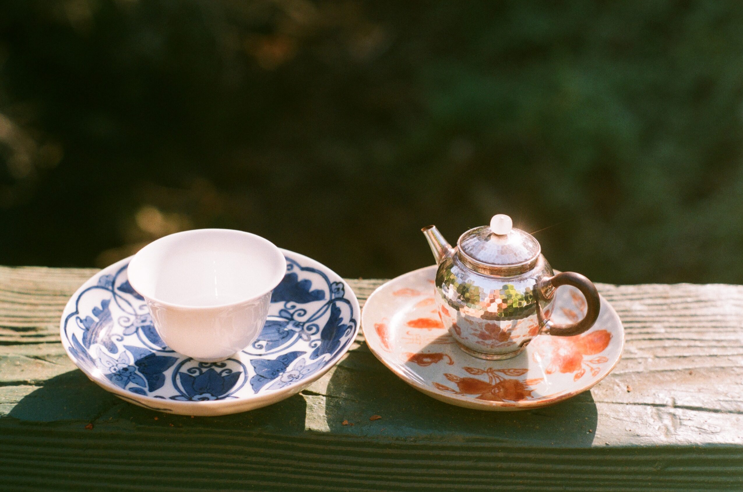 Tea set with a teapot, cup, and saucer on a wooden surface outdoors, with blurred greenery in the background.