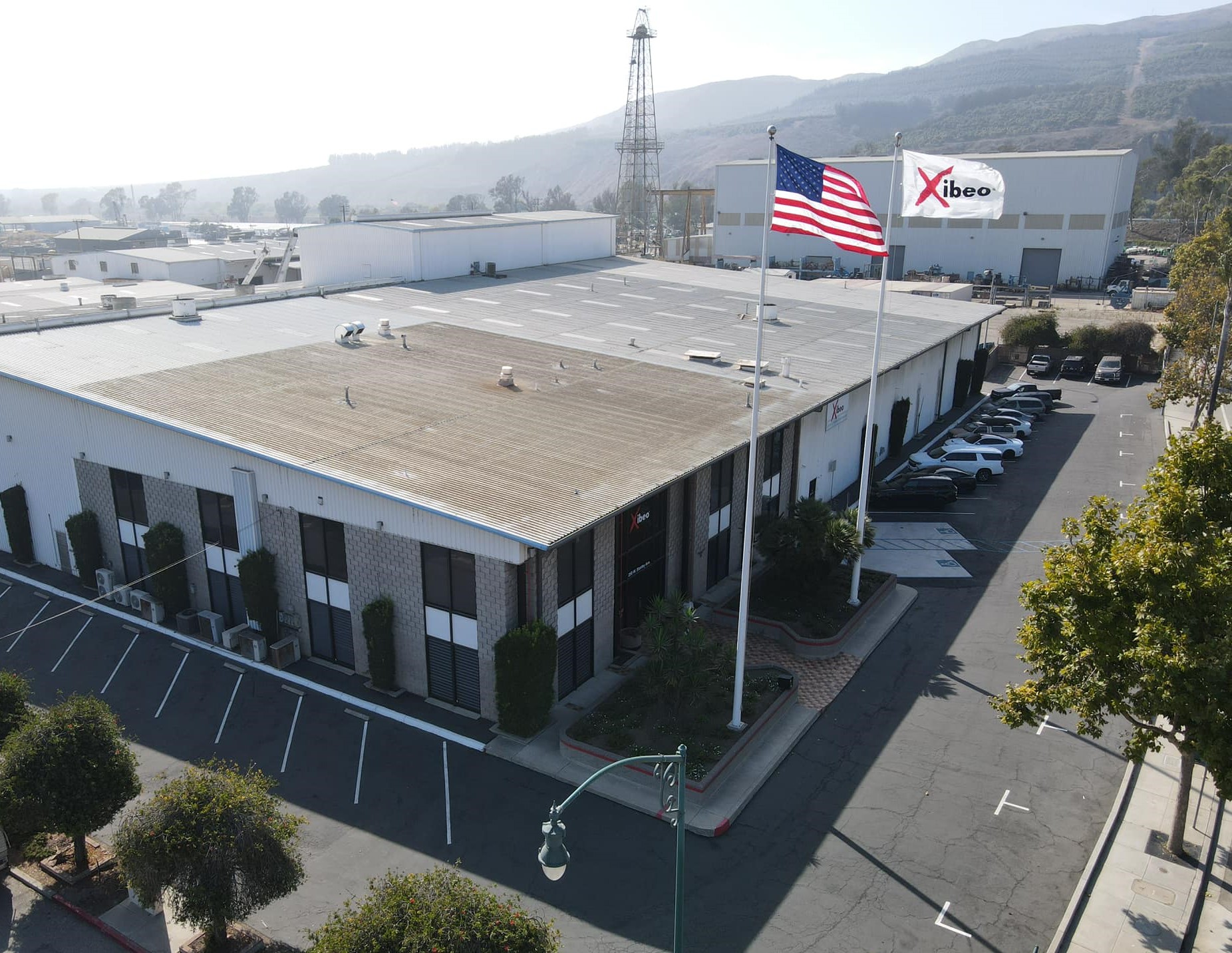 A large industrial building with parking lot, American flag, and company sign, surrounded by trees and mountains in background.