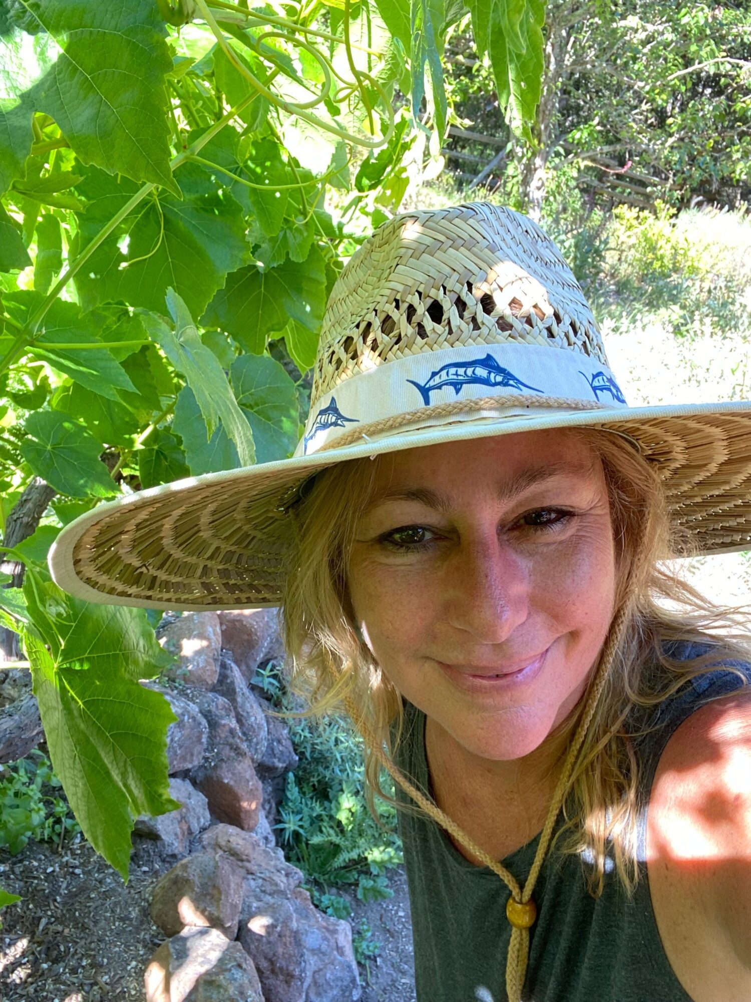 Woman wearing a wide-brimmed hat outdoors among green plants and rocks, smiling at the camera.