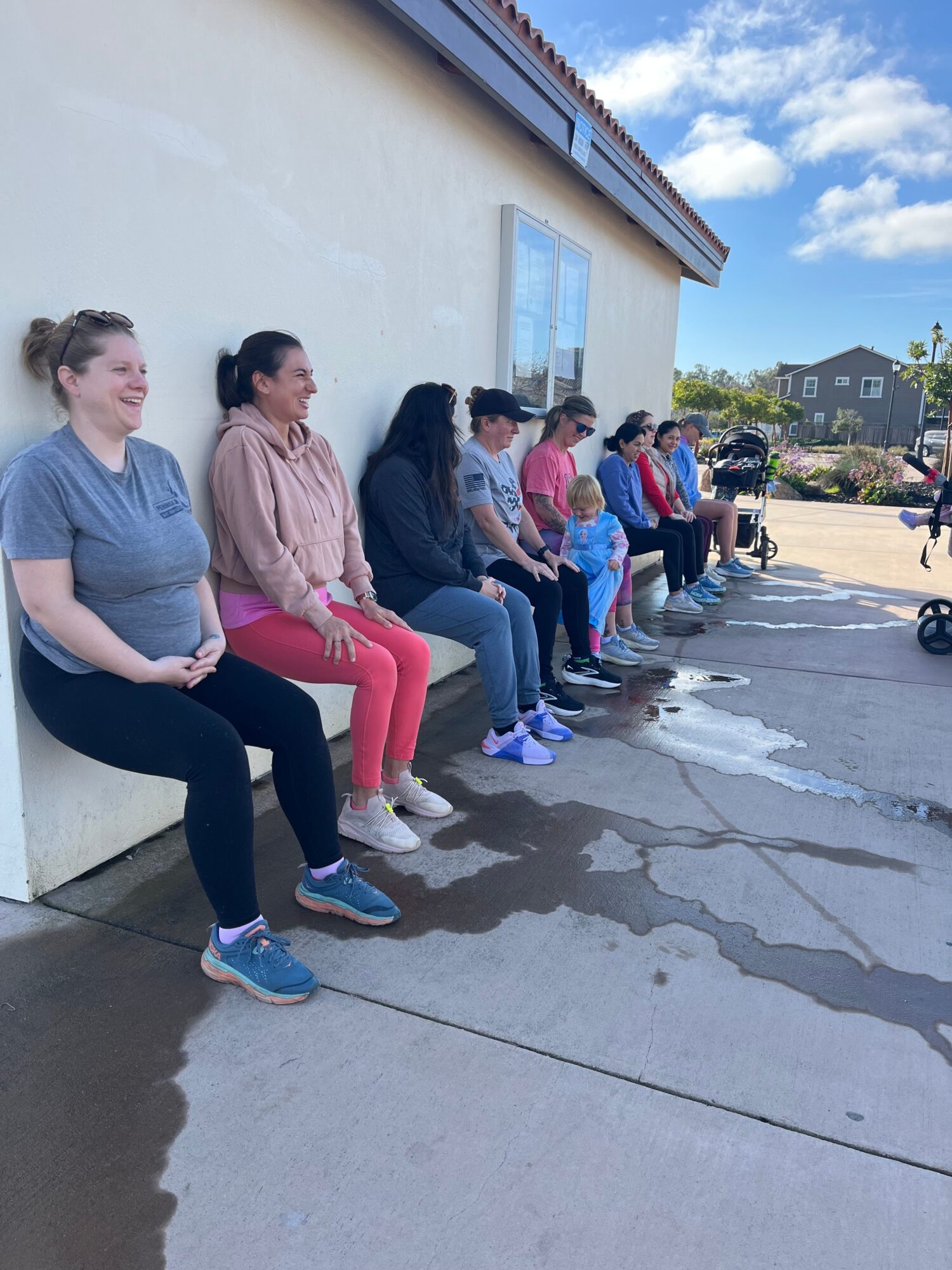 Group of people sitting on a sidewalk outside a building, some with water puddles nearby, under a partly cloudy sky.