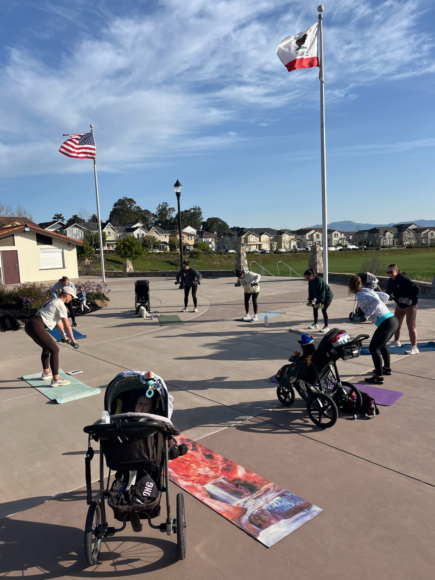 Group of people practicing yoga outdoors on a sunny day, with flags and a stroller nearby, in a park-like setting.