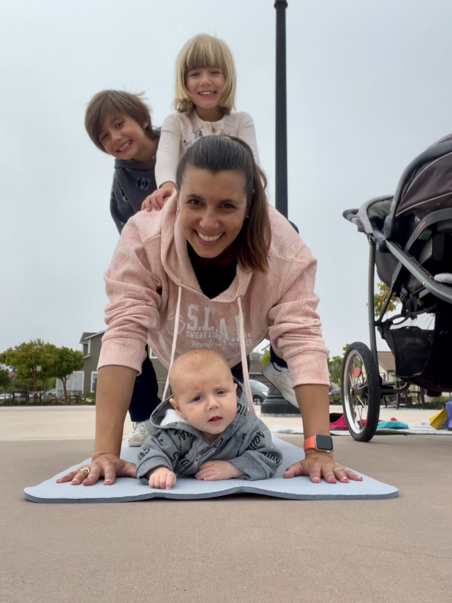 Woman doing a plank with two children on her back and one baby on the ground, outdoors on a cloudy day.