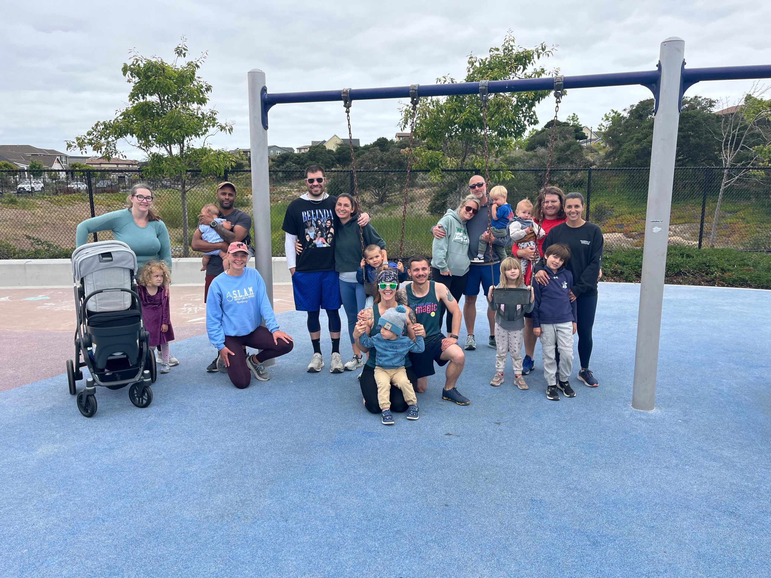 Group of people, including children, adults, and a person with a stroller, standing on a blue playground surface near a swing set, outdoors.