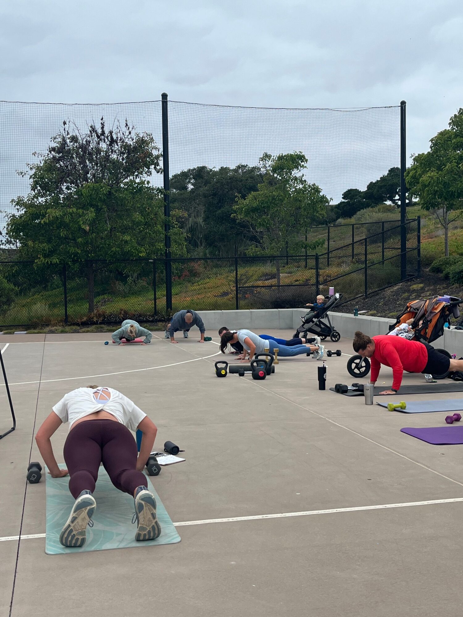People exercising on a concrete outdoor court with trees and a fence in the background, some using mats and weights.