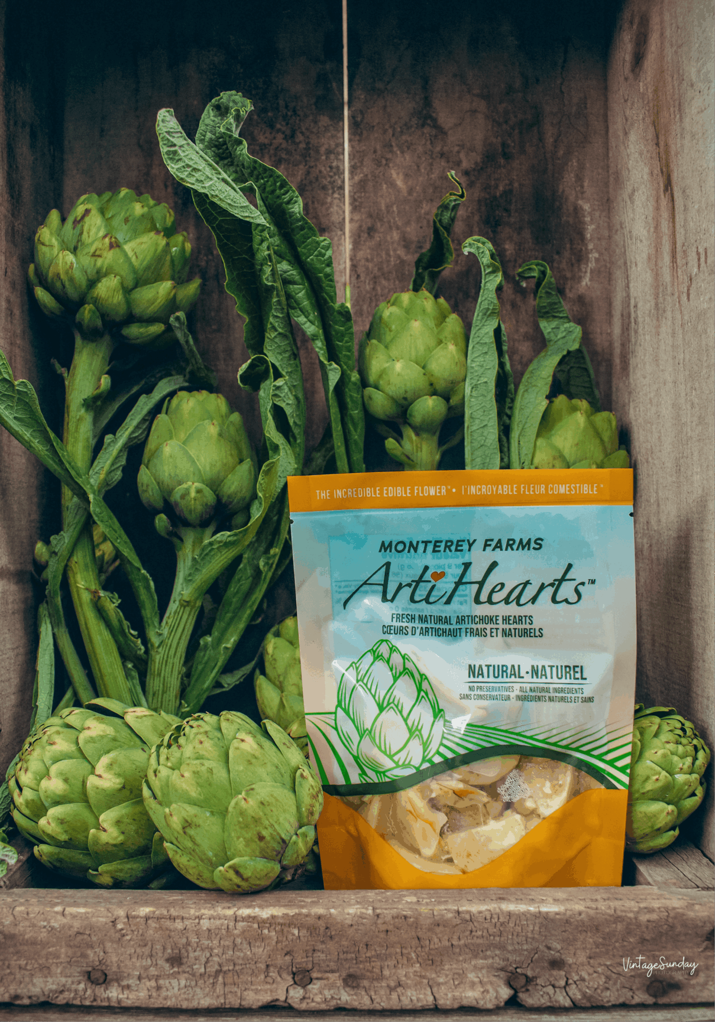Green artichokes with leaves behind a bag of Monterey Farms Art Hearts snacks on a wooden surface.