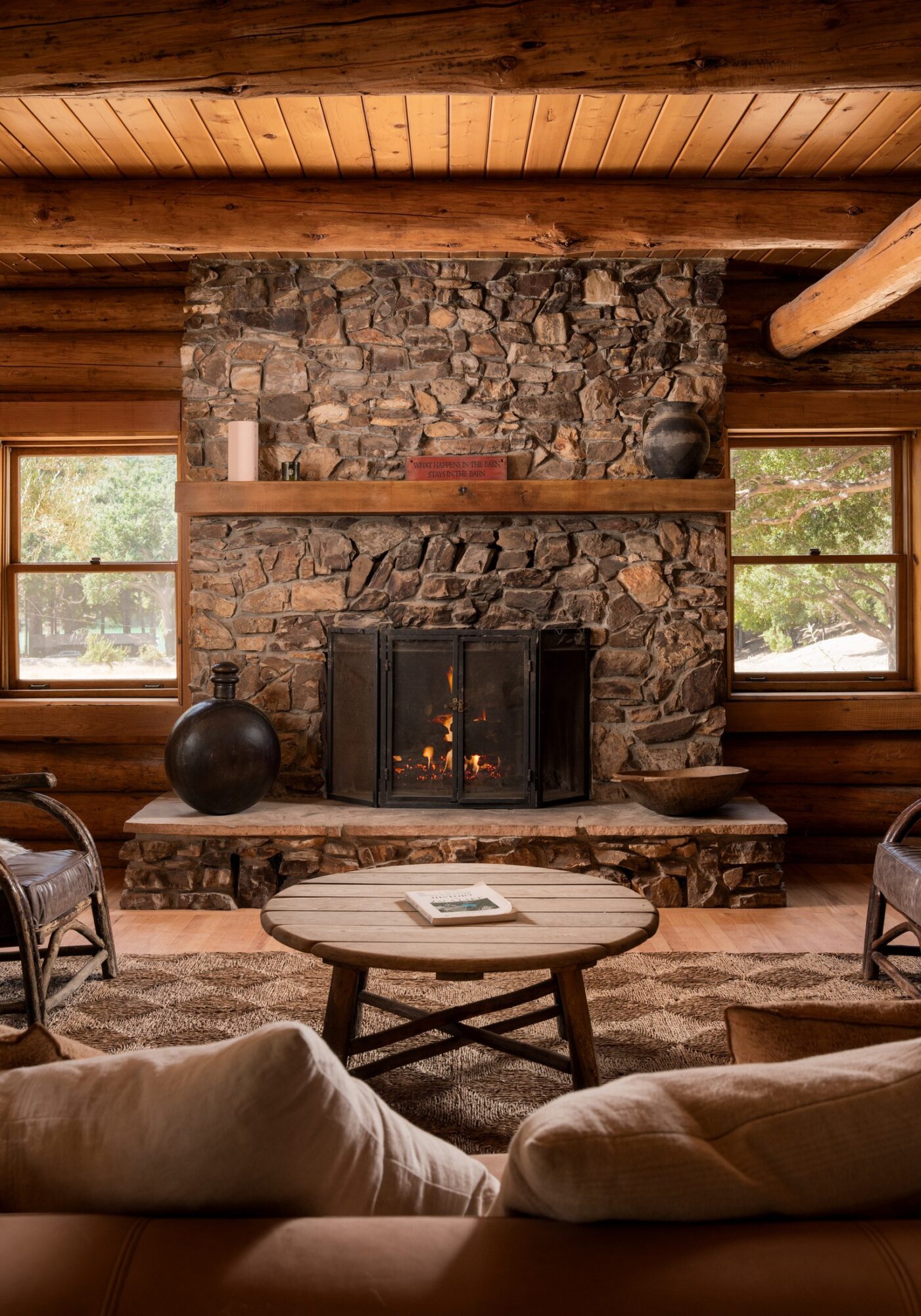 Living room with stone fireplace, wooden ceiling, windows, and cozy seating around a round table.