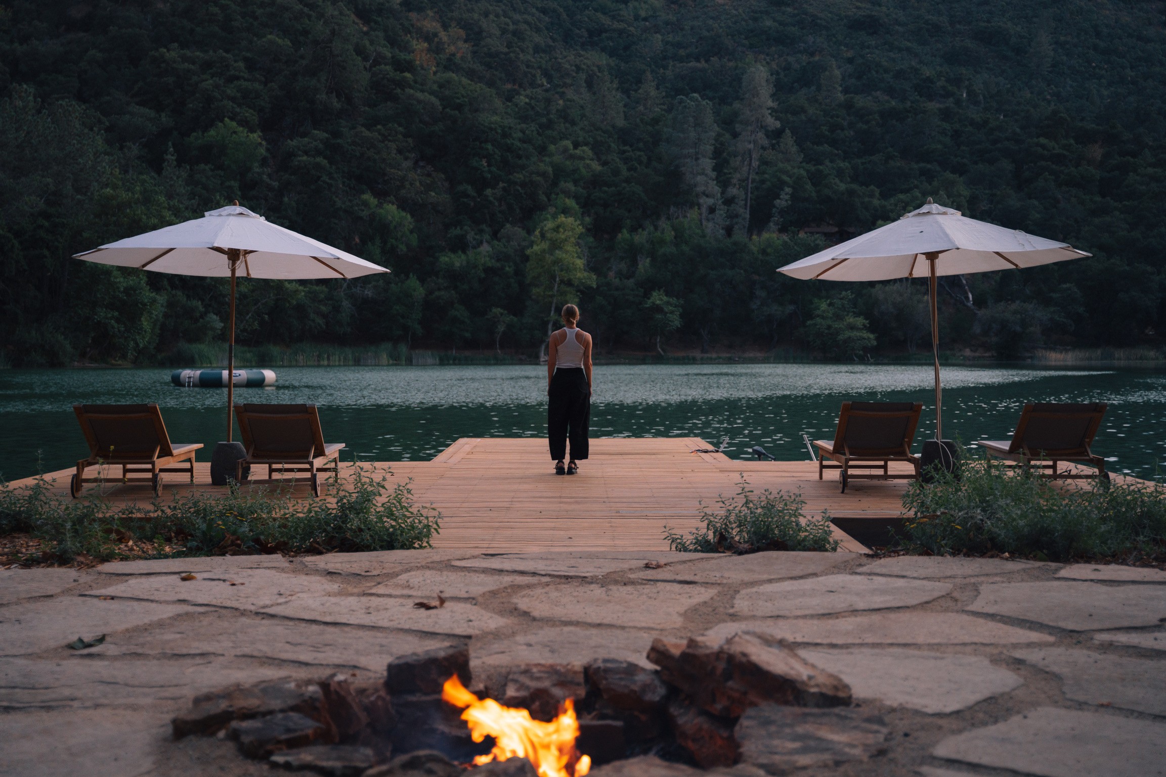 Person standing on dock by lake with two umbrellas and lounge chairs, surrounded by trees and mountains.