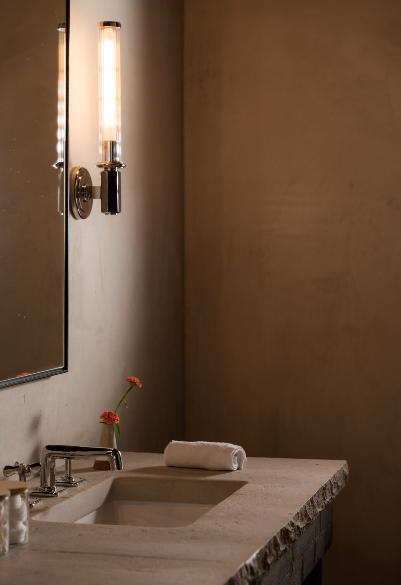 Bathroom sink with a rolled towel and a small flower in a vase, illuminated by a wall-mounted light fixture.