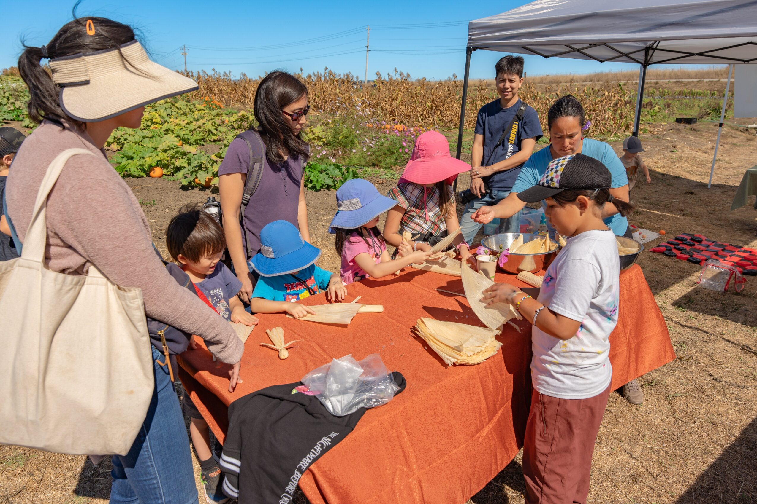 Group of children and adults gathered around a table outdoors, engaging in an activity under a canopy, with a field in the background.