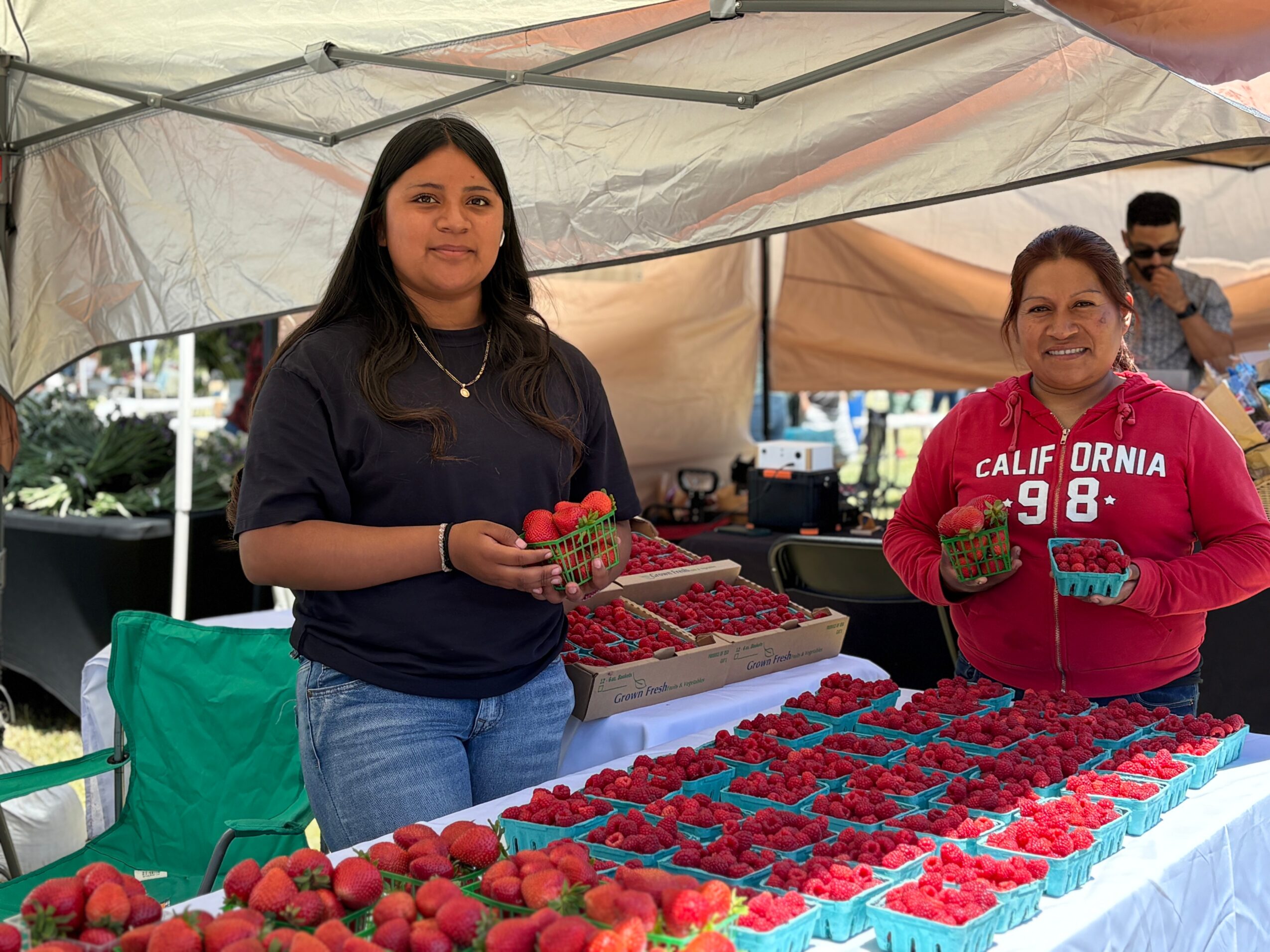 Two women stand behind a table with strawberries and berries at an outdoor market stall under a canopy.