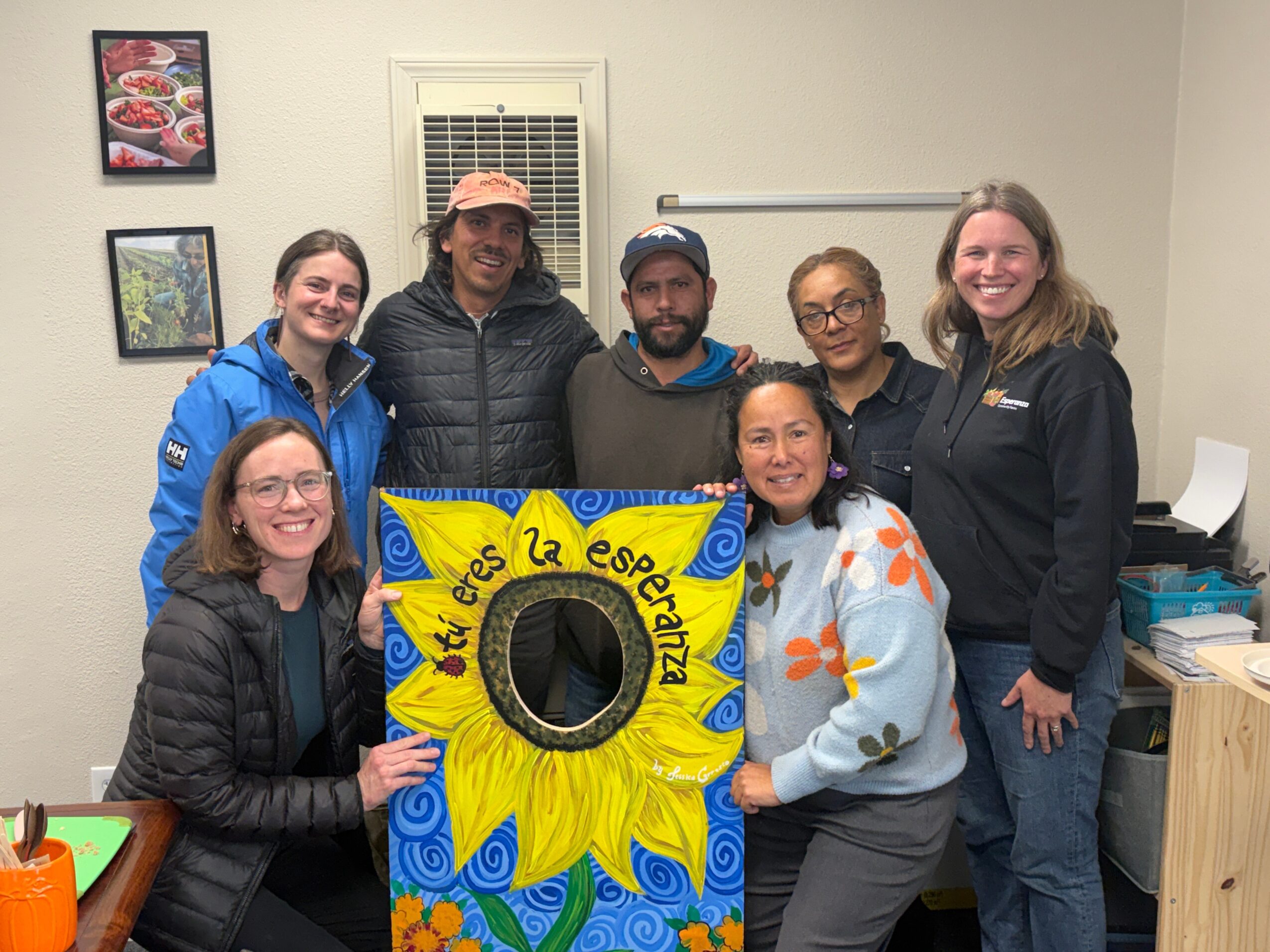 Group of seven people holding a colorful sunflower-themed sign indoors, smiling, with framed pictures on the wall behind.