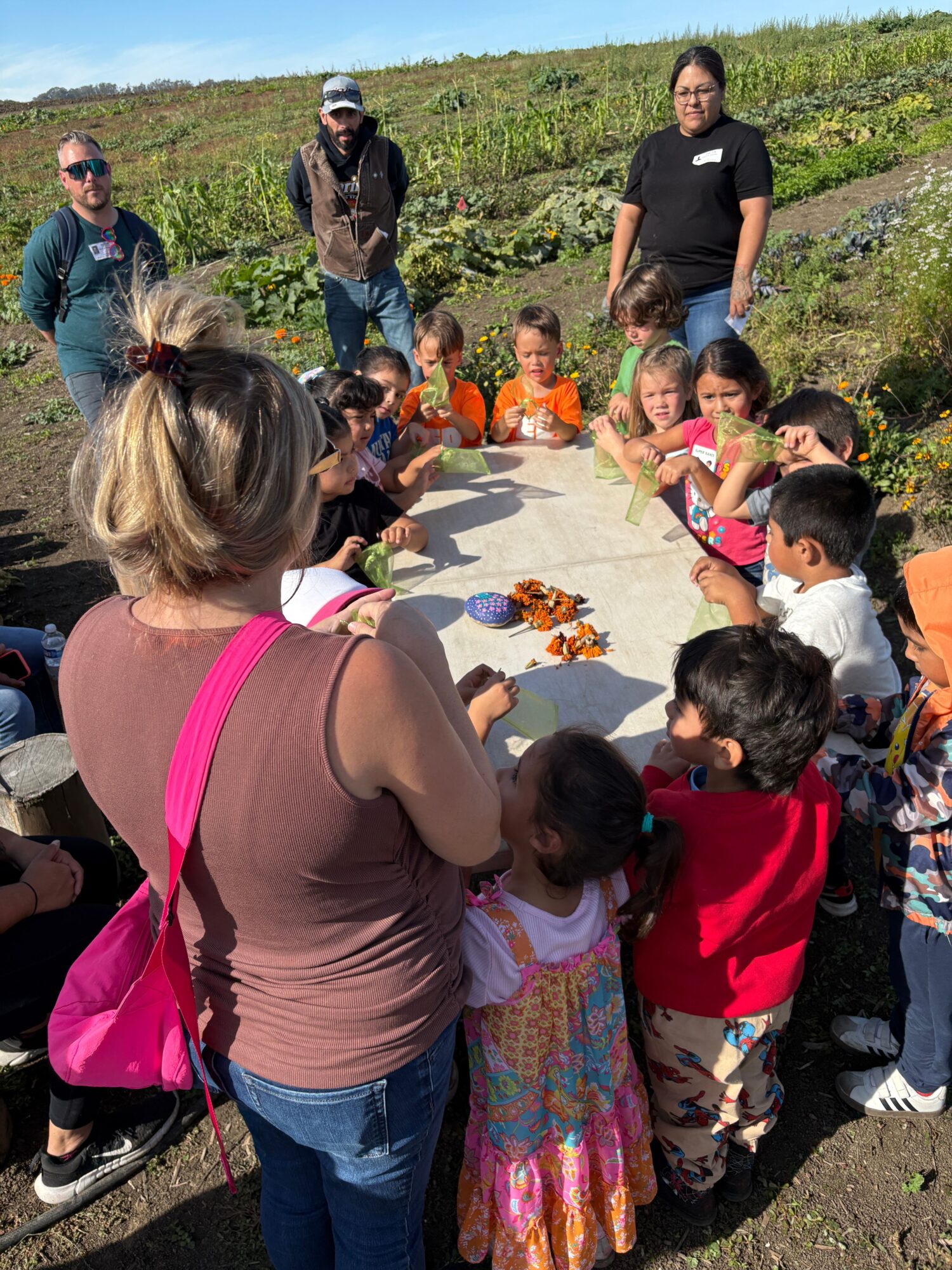 Group of children and adults gathered outdoors around a table with decorated butterflies, on a sunny day.