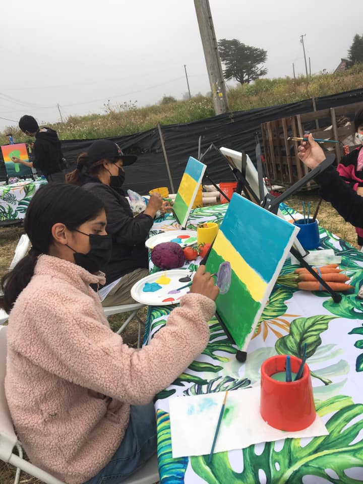 People painting on canvases outdoors at a table with a colorful tablecloth, trees, and a fence in the background.