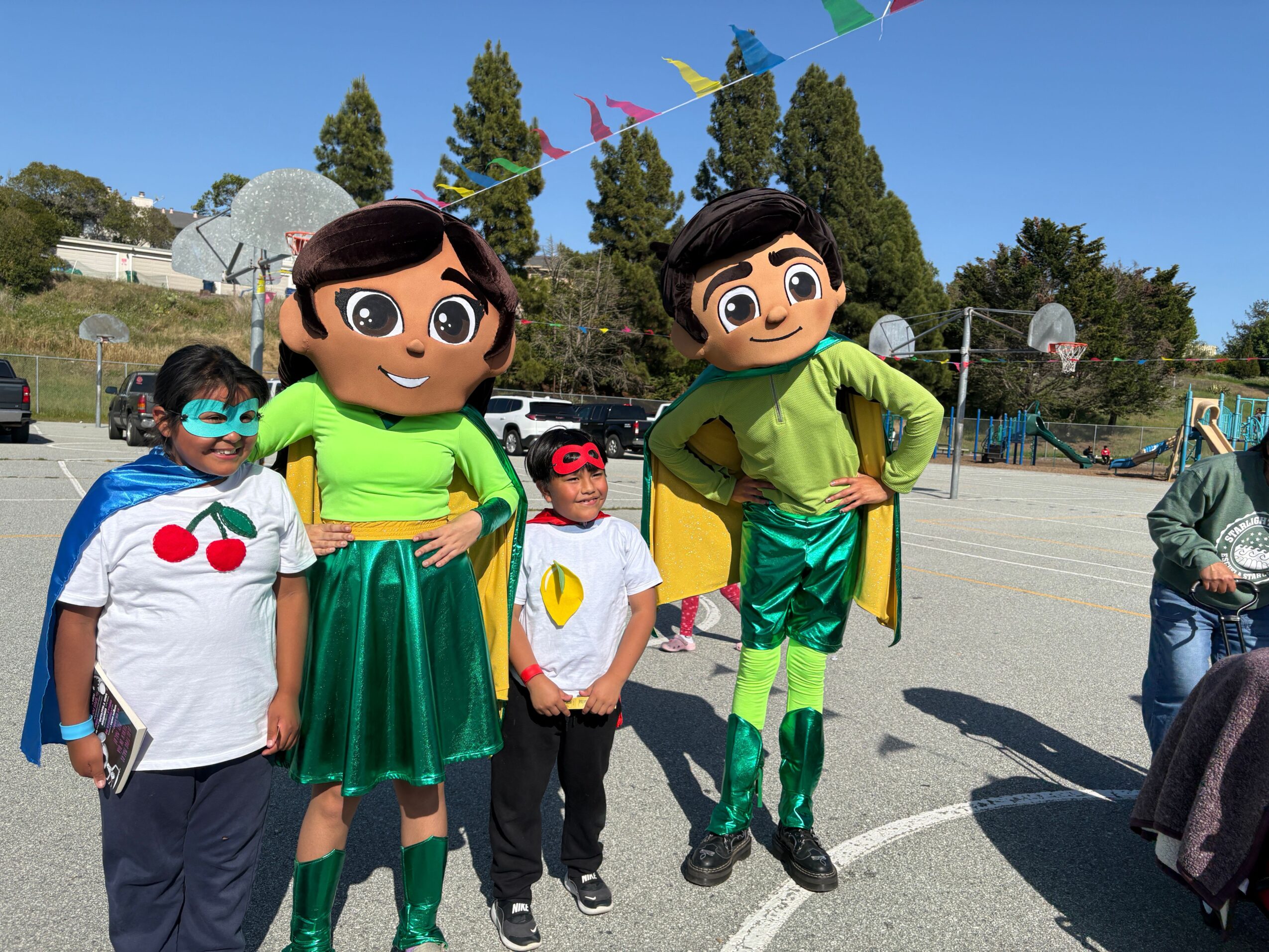 Group of children in costumes standing outdoors on a sunny day, with trees and a road in the background.