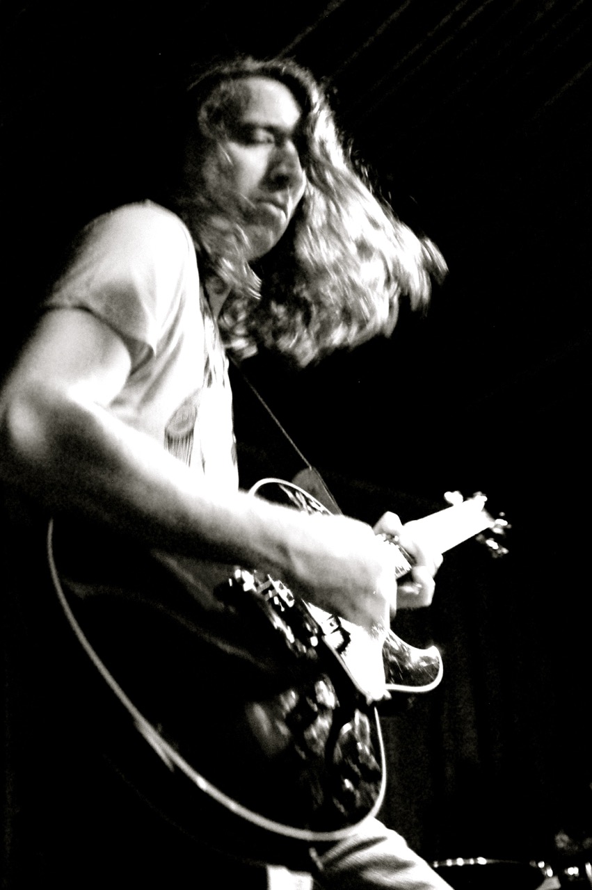 Person with long hair playing an electric guitar on stage, black and white photo, focused expression.