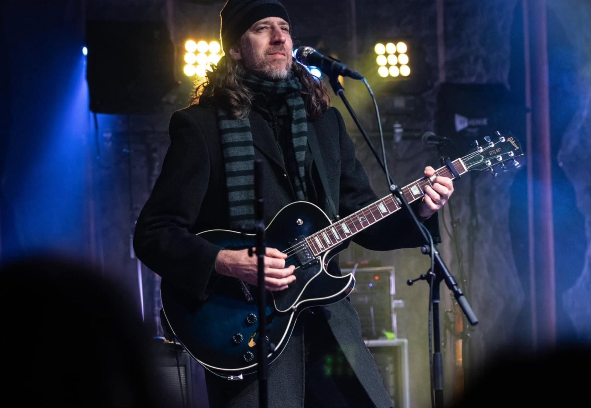 Man with long hair and beard playing an acoustic guitar on stage with bright lights behind him.