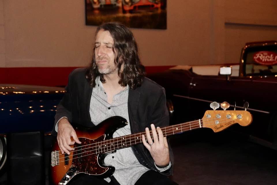 Person with long hair playing a bass guitar in a room with a red and black background.