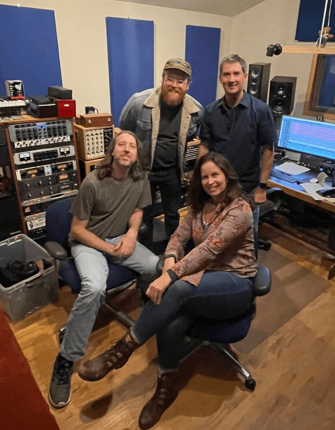 Four people in a recording studio, two seated and two standing, surrounded by audio equipment and a computer screen.