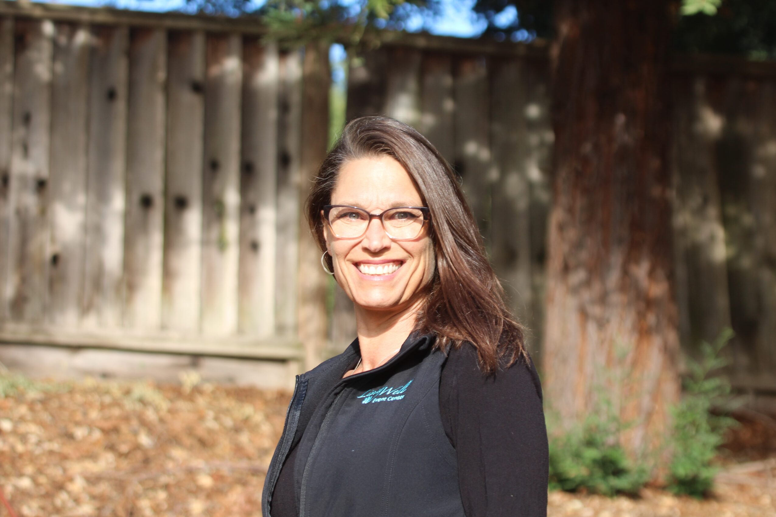 Woman with glasses smiling outdoors, wearing a black jacket, in front of a wooden fence and trees.