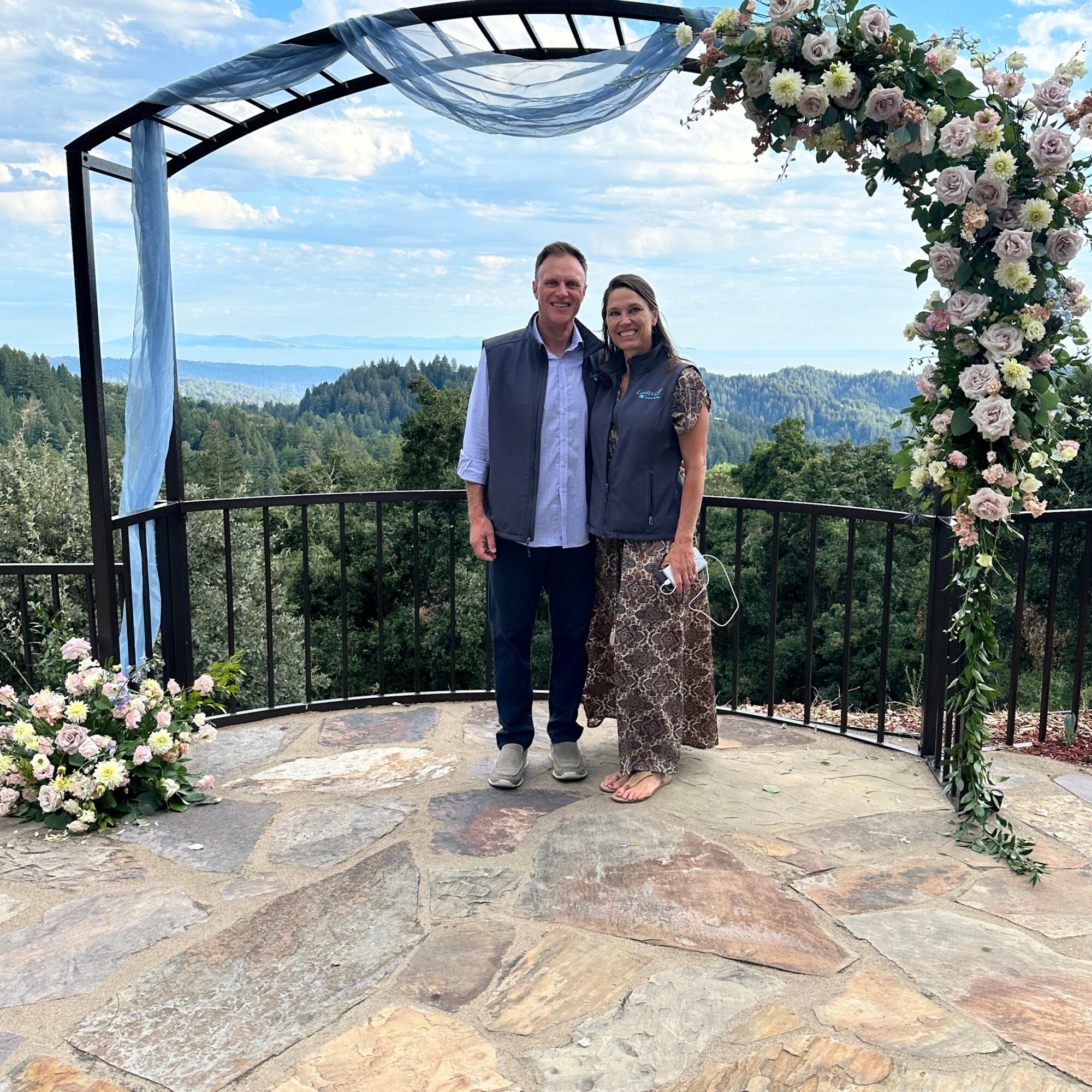 Two people standing under a decorated arch on a balcony with a scenic landscape background.