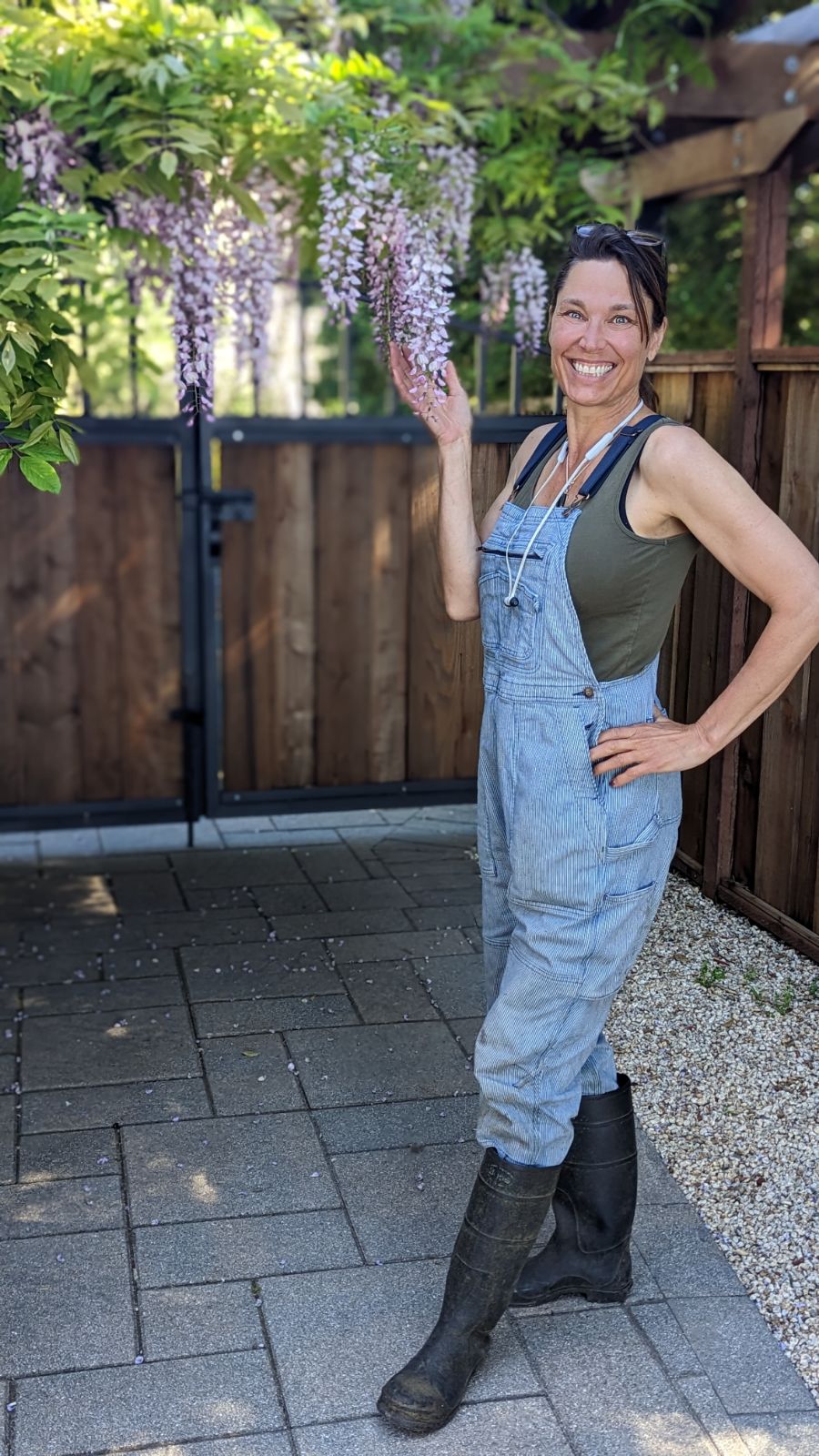 Woman in overalls and boots standing outdoors near a wooden fence, smiling, with hanging purple flowers above her.