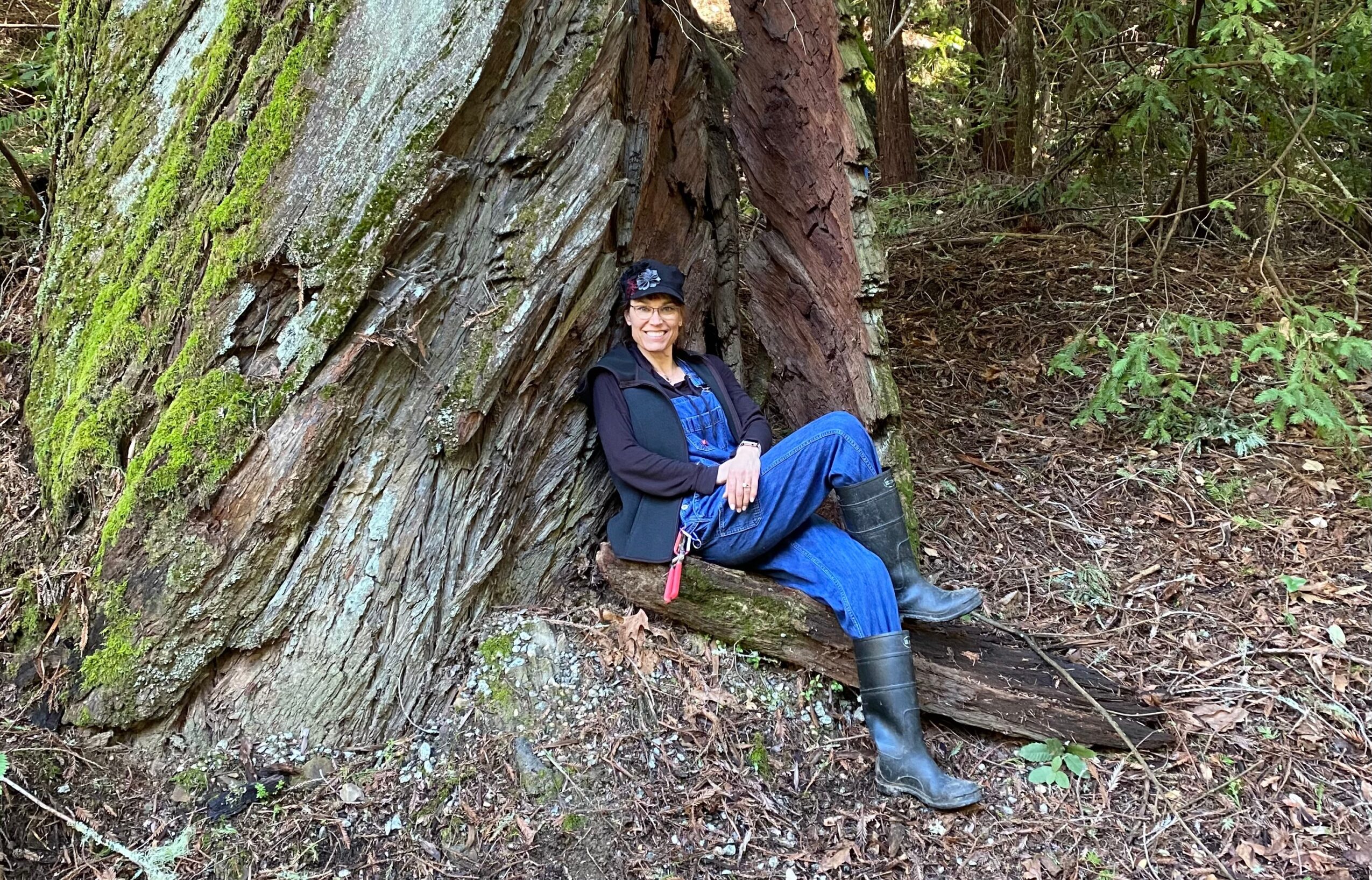 Person sitting against a large moss-covered tree trunk in a forest, smiling, wearing a hat, jacket, and boots.