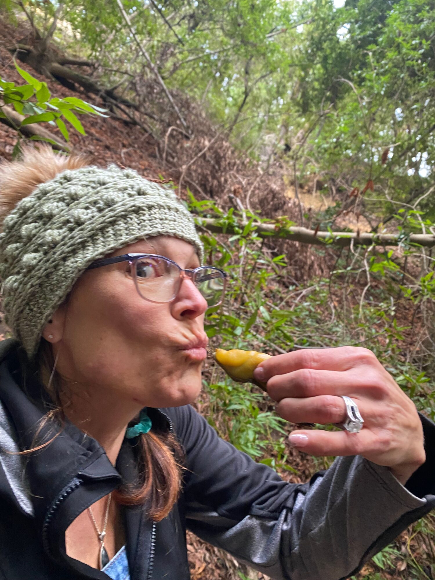 Person with glasses and a knitted hat in a forest, holding a yellow object near their mouth.