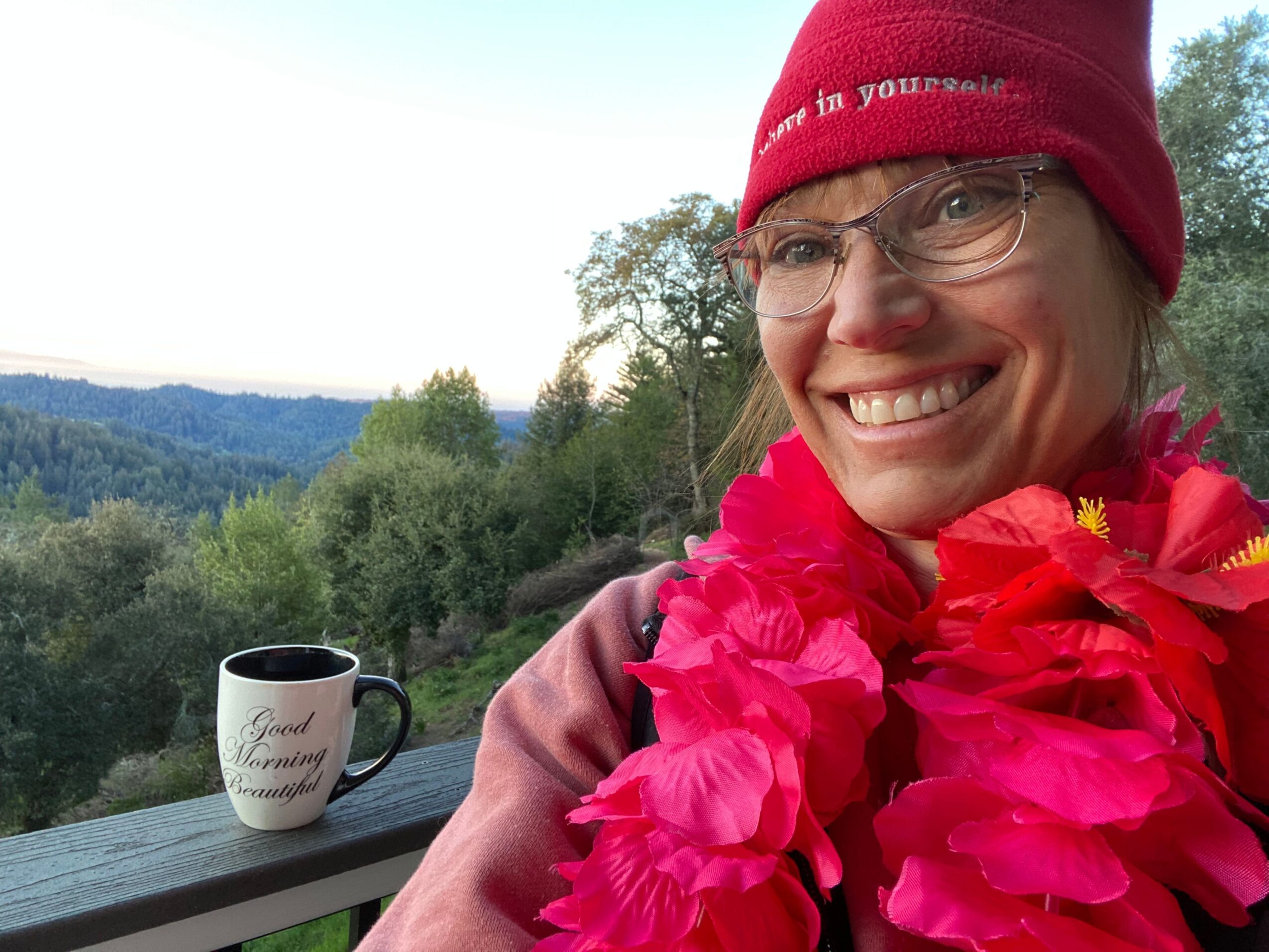 Smiling woman wearing glasses and a red hat, holding pink flowers, outdoors with trees and a lake in the background.