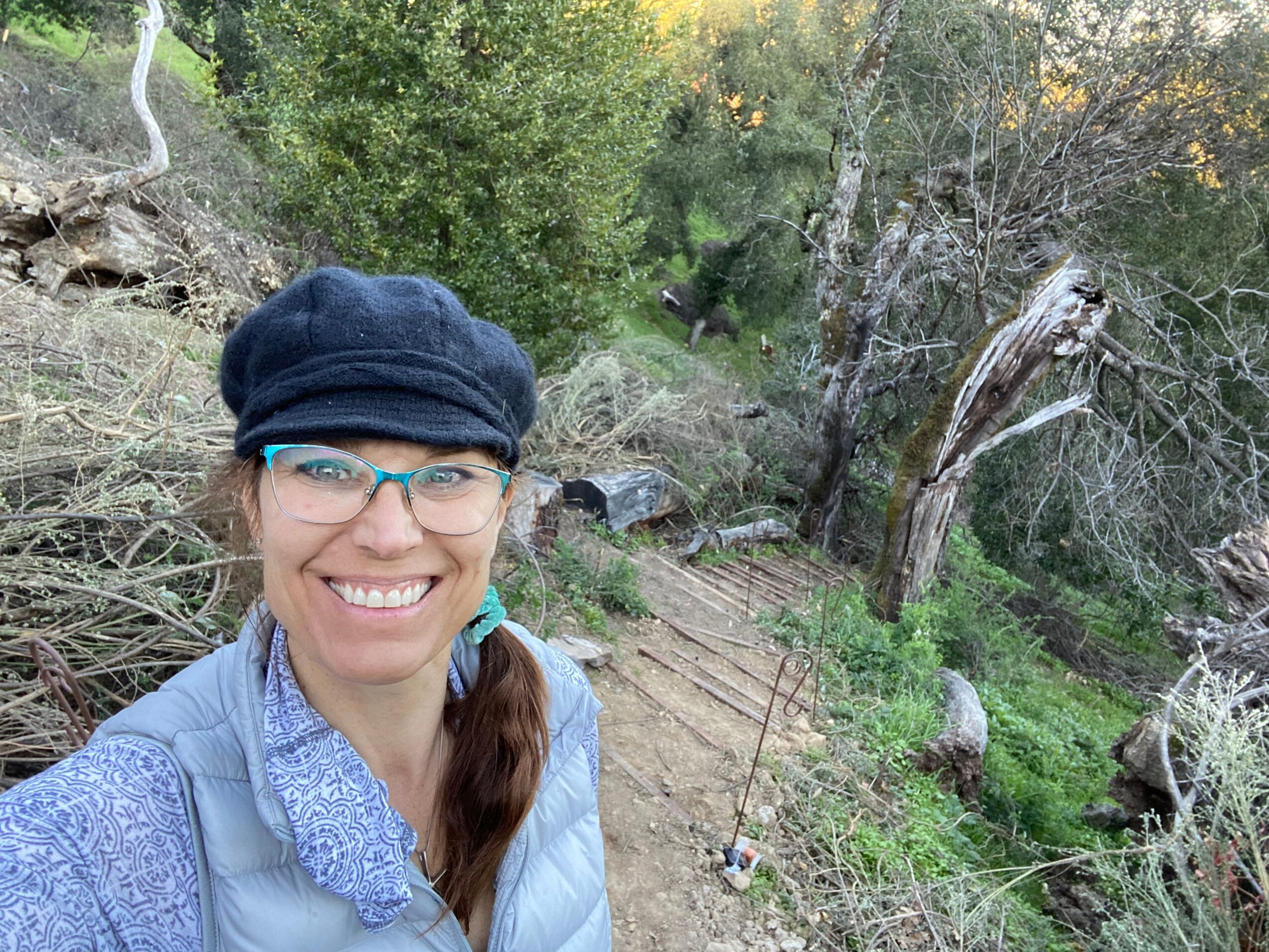 Person smiling outdoors in a wooded area, wearing glasses, a blue jacket, and a dark hat, with trees and greenery around.
