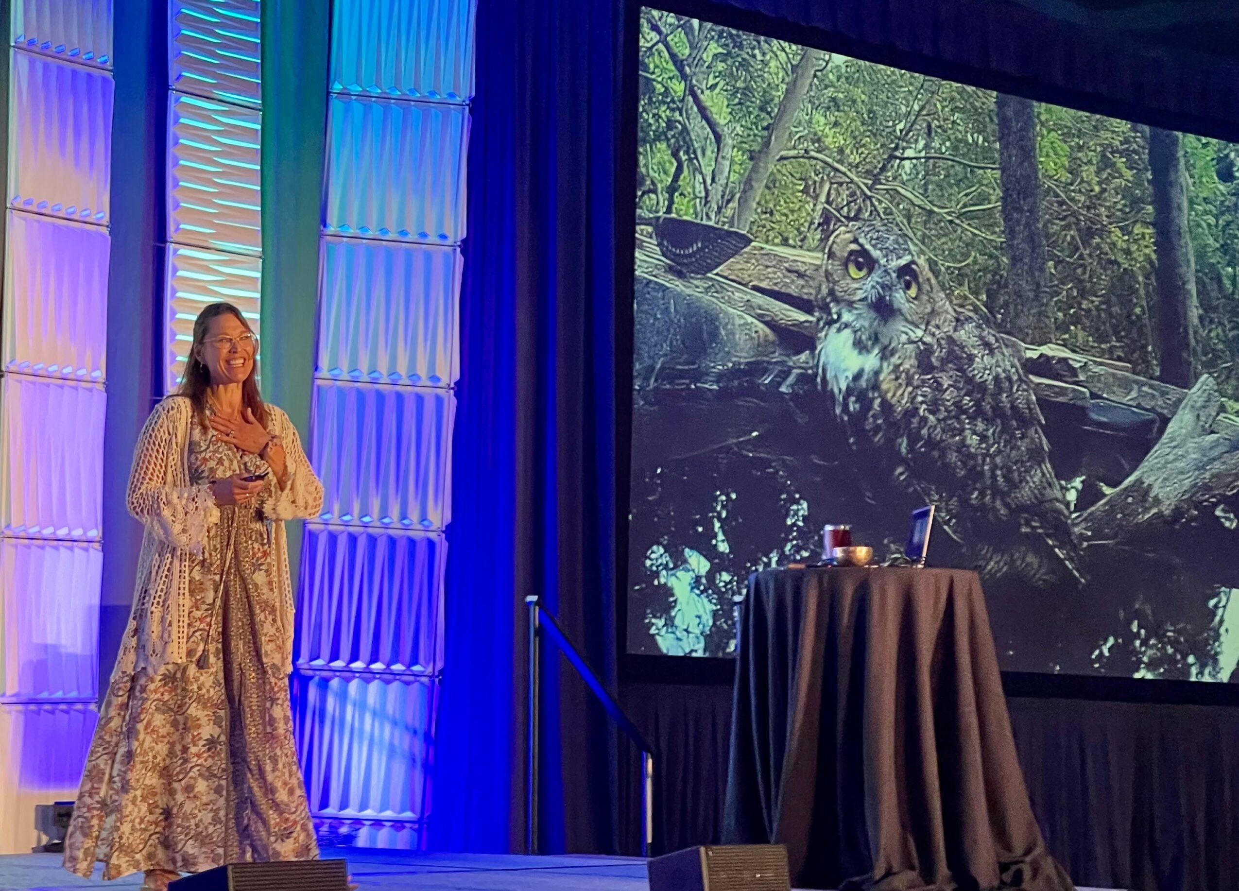Woman on stage with colorful background, large screen showing an owl in a forest, and a table with items.