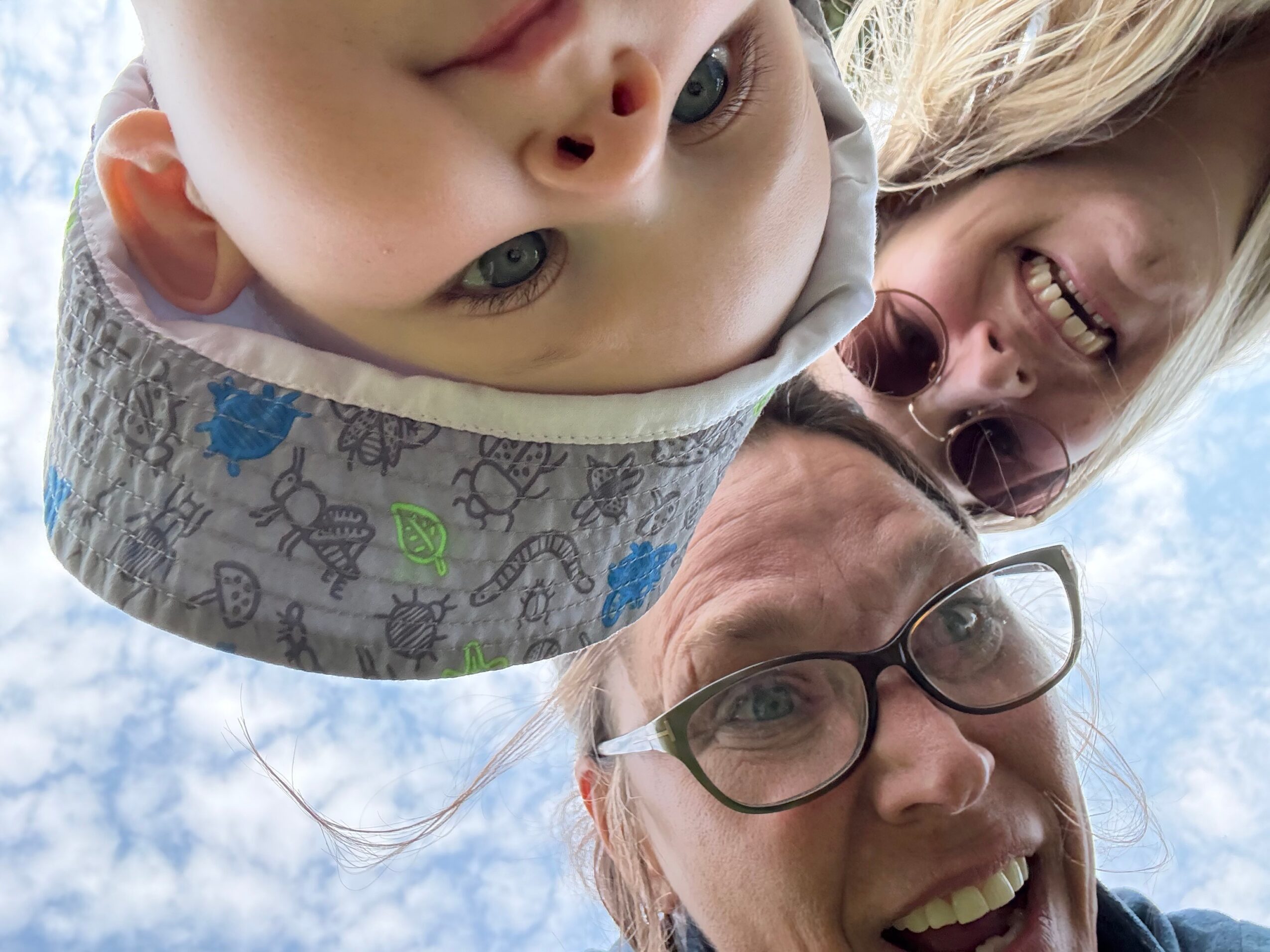 Family of three smiling, upside down, outdoors with blue sky and clouds, close-up view of faces.