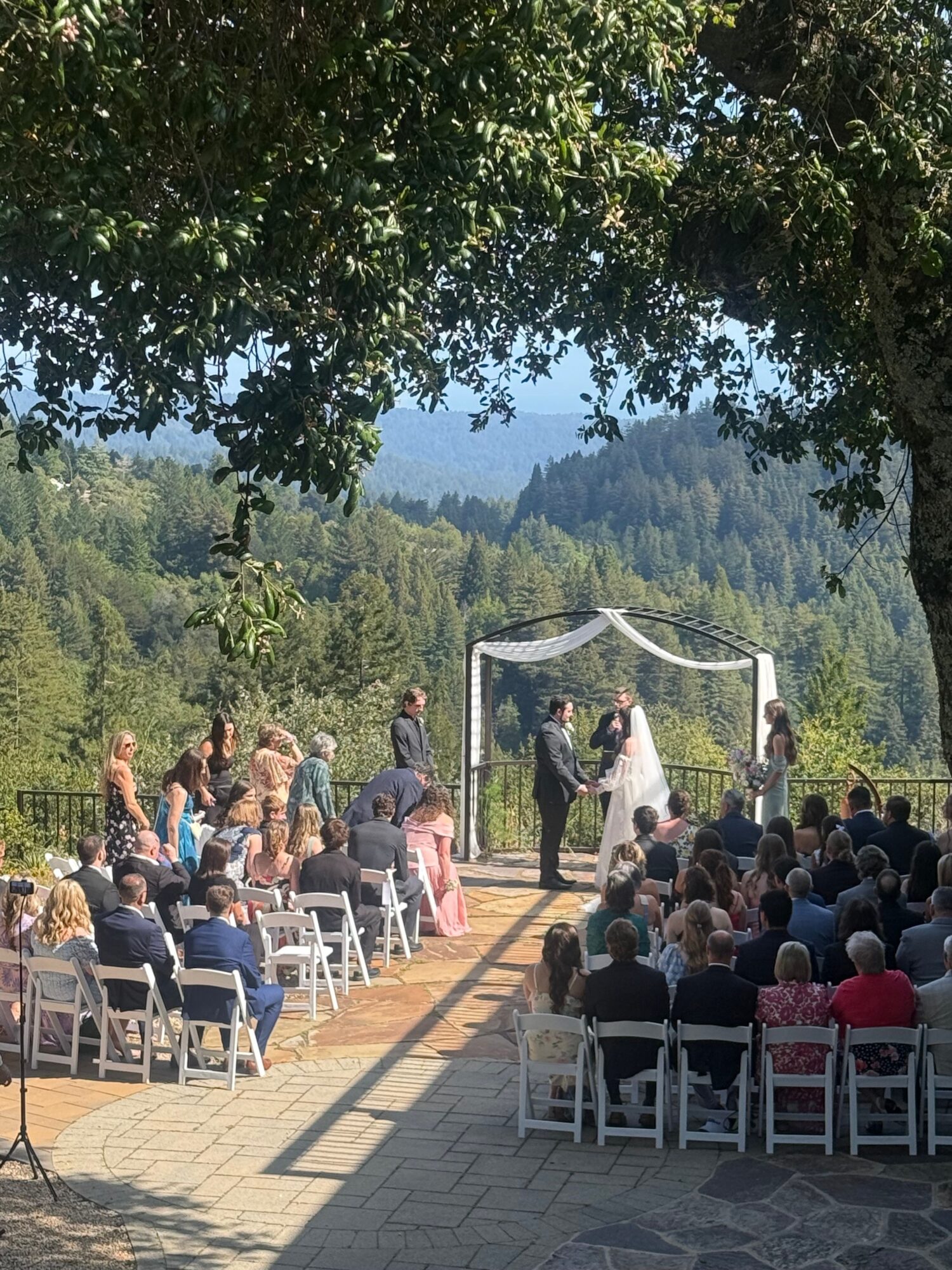 People gathered outdoors on chairs facing a speaker at a wooden stage with a scenic mountain background.