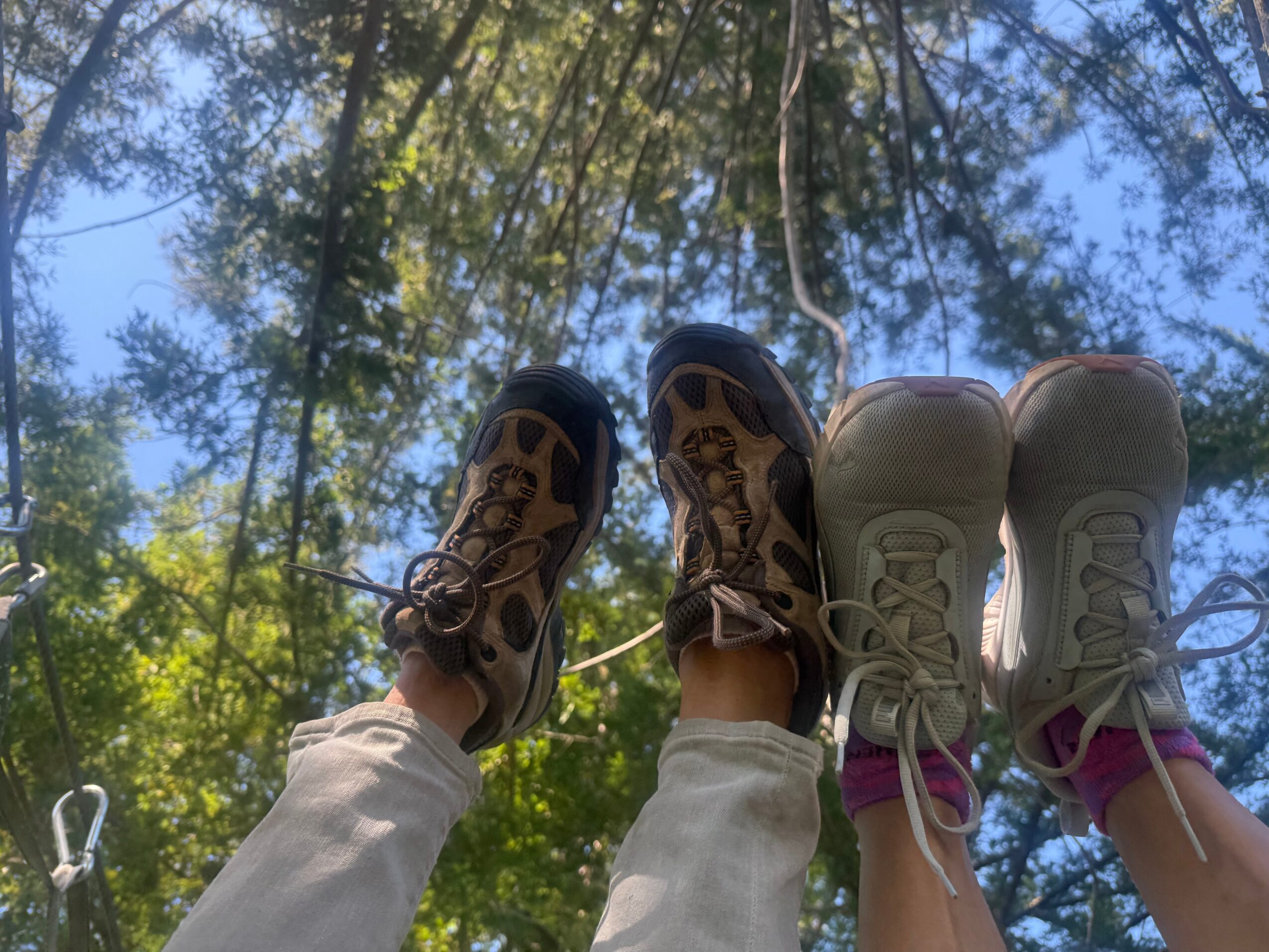 Two people standing outdoors on a sunny day, with trees and blue sky overhead, wearing sneakers and light-colored pants.