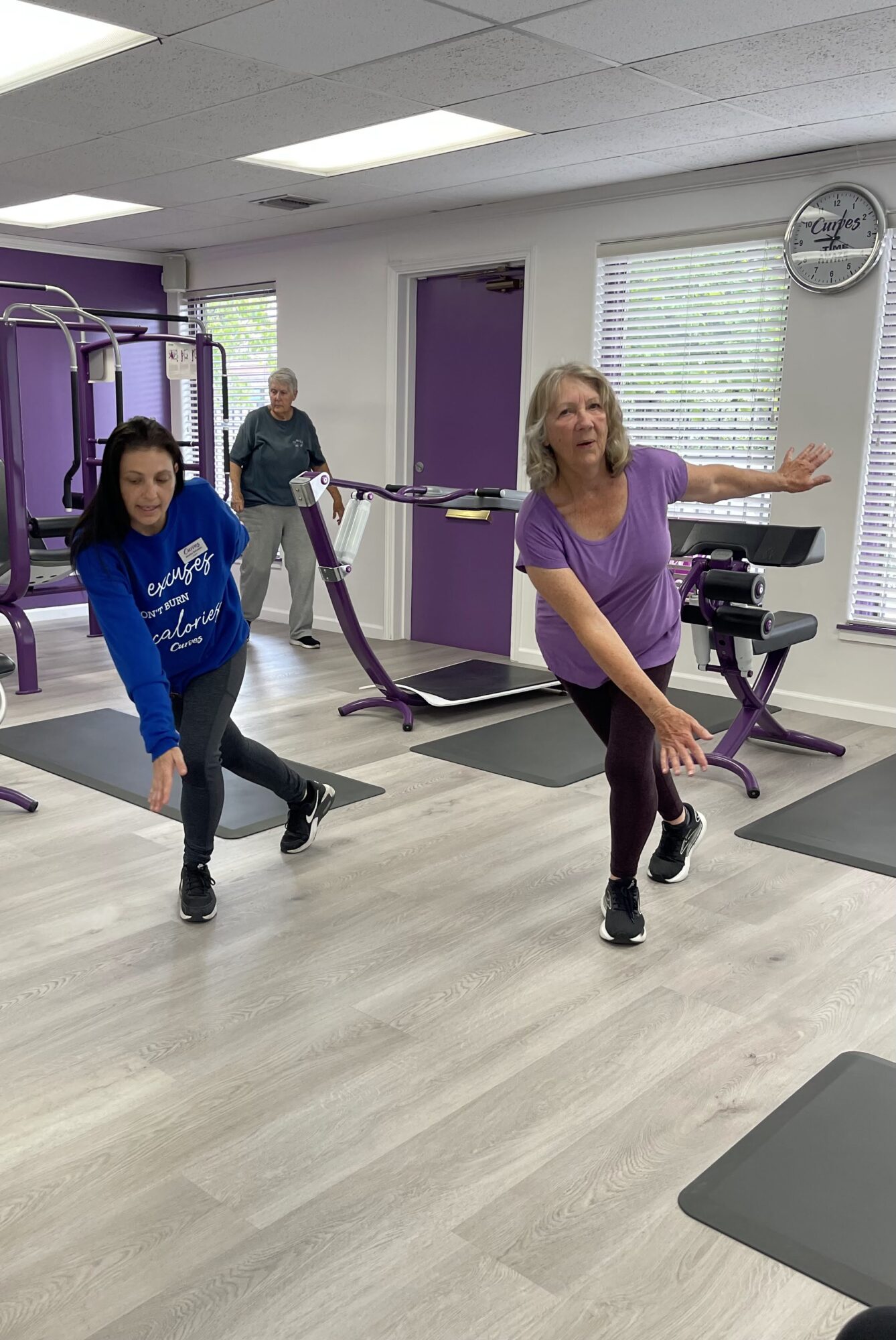 Three women exercising in a gym with purple and white walls, gym equipment, and windows with blinds.