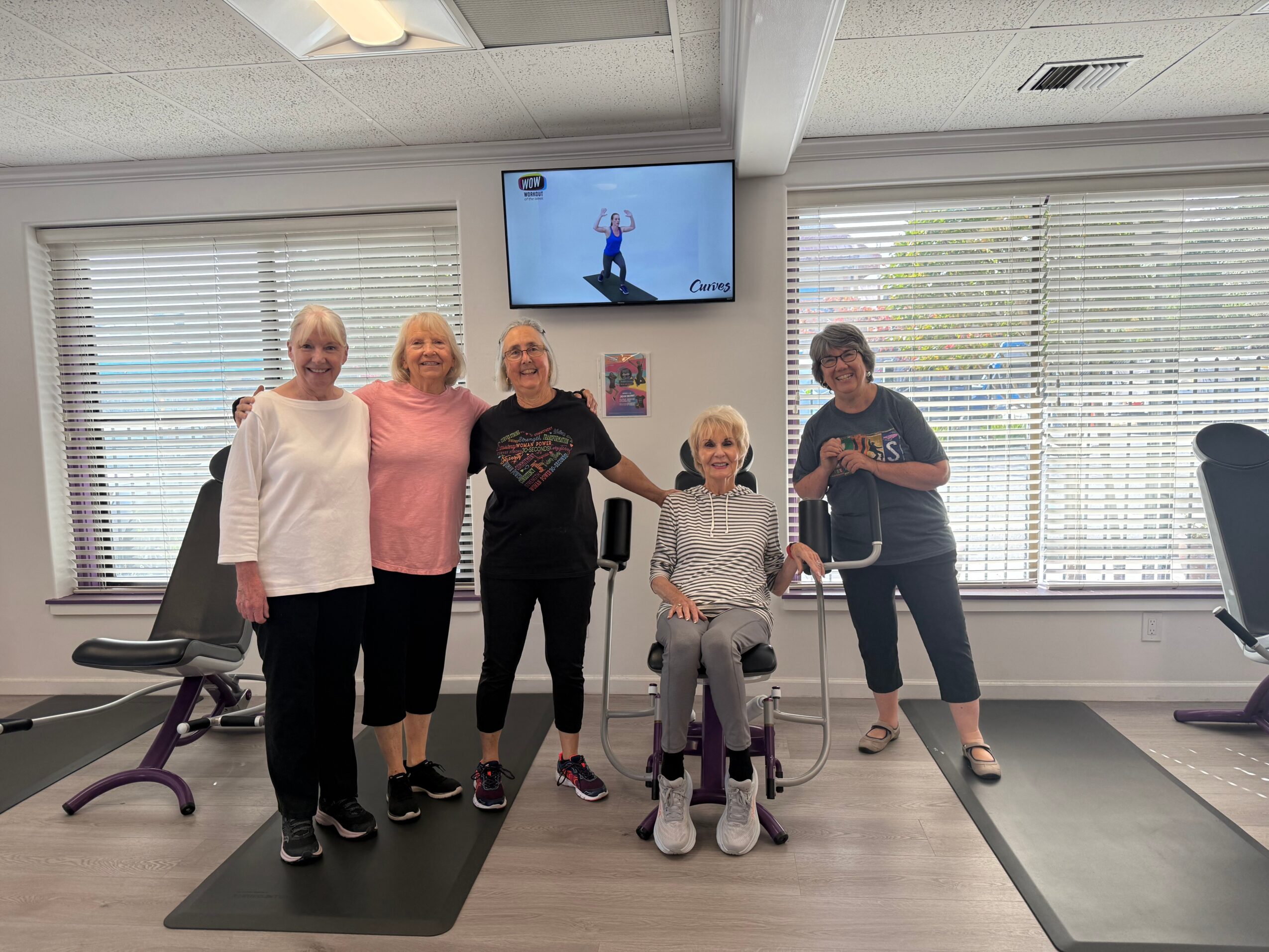 Five women standing in a gym, one seated in a wheelchair, smiling, with exercise equipment and windows in the background.