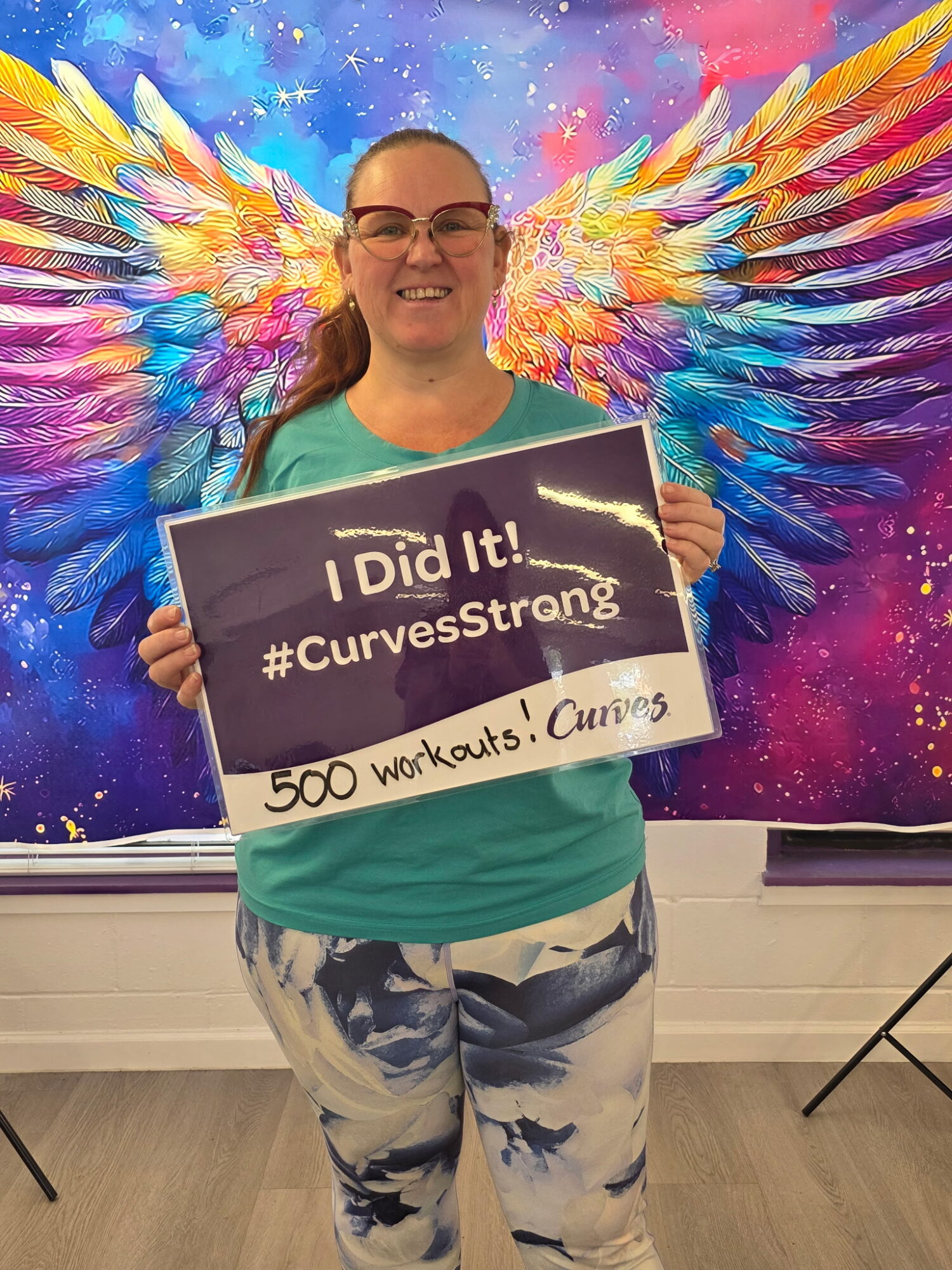 Smiling woman holding a sign with text, standing in front of colorful angel wings mural.