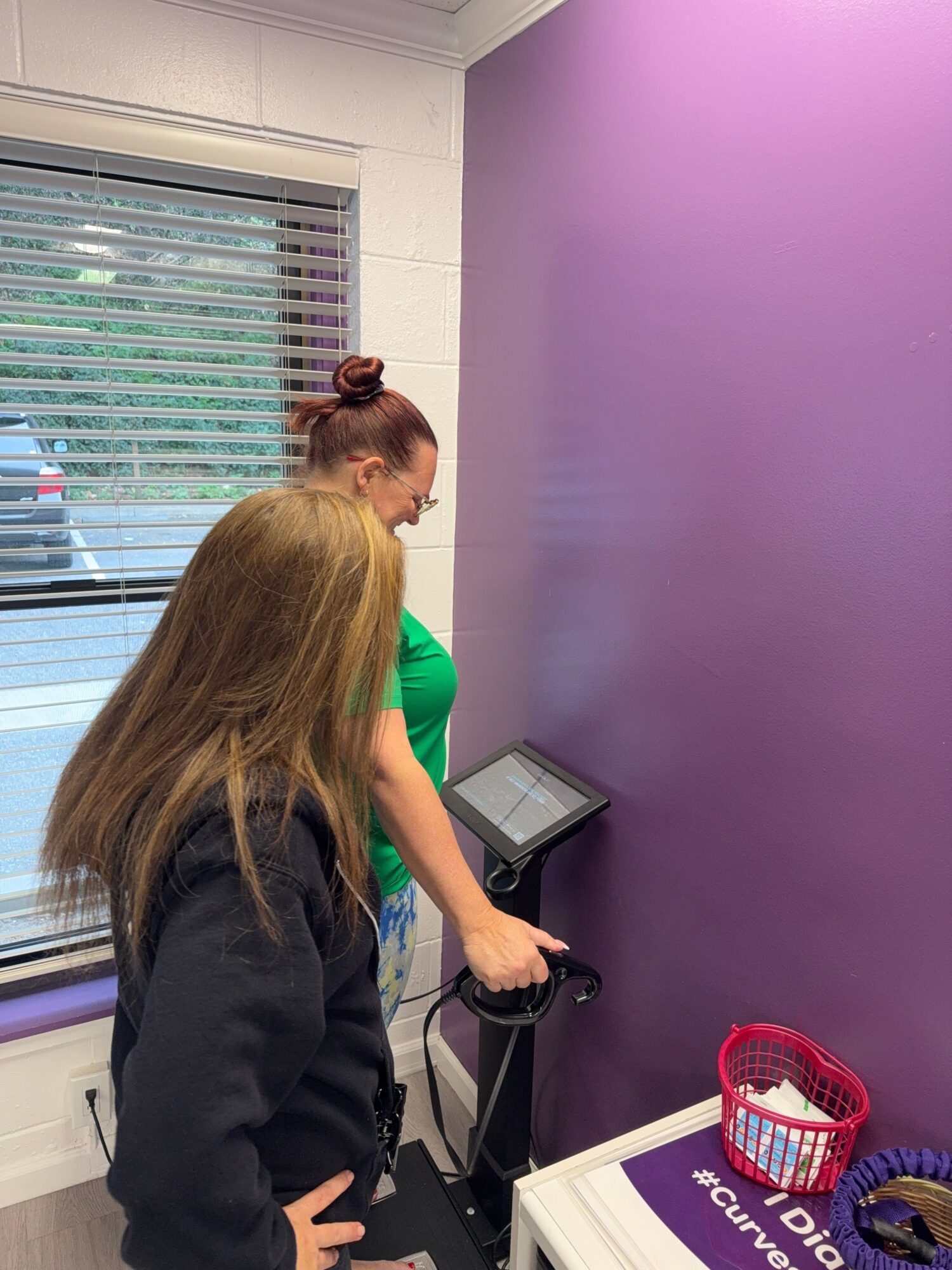 Two women in a room with purple walls, one using a device, the other observing, near a window with blinds.