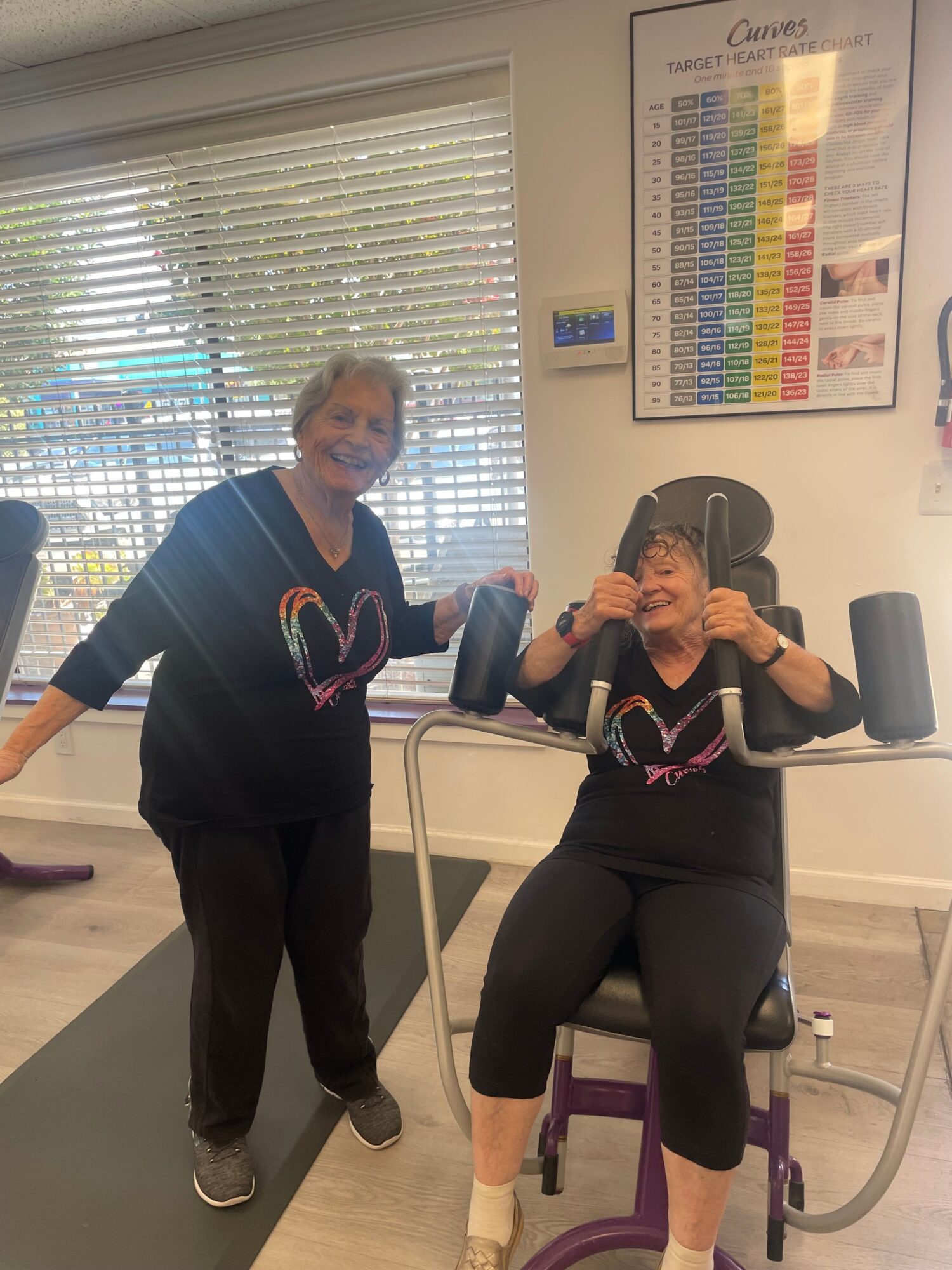 Two women in black workout clothes sitting on exercise machines in a gym, smiling and holding water bottles.