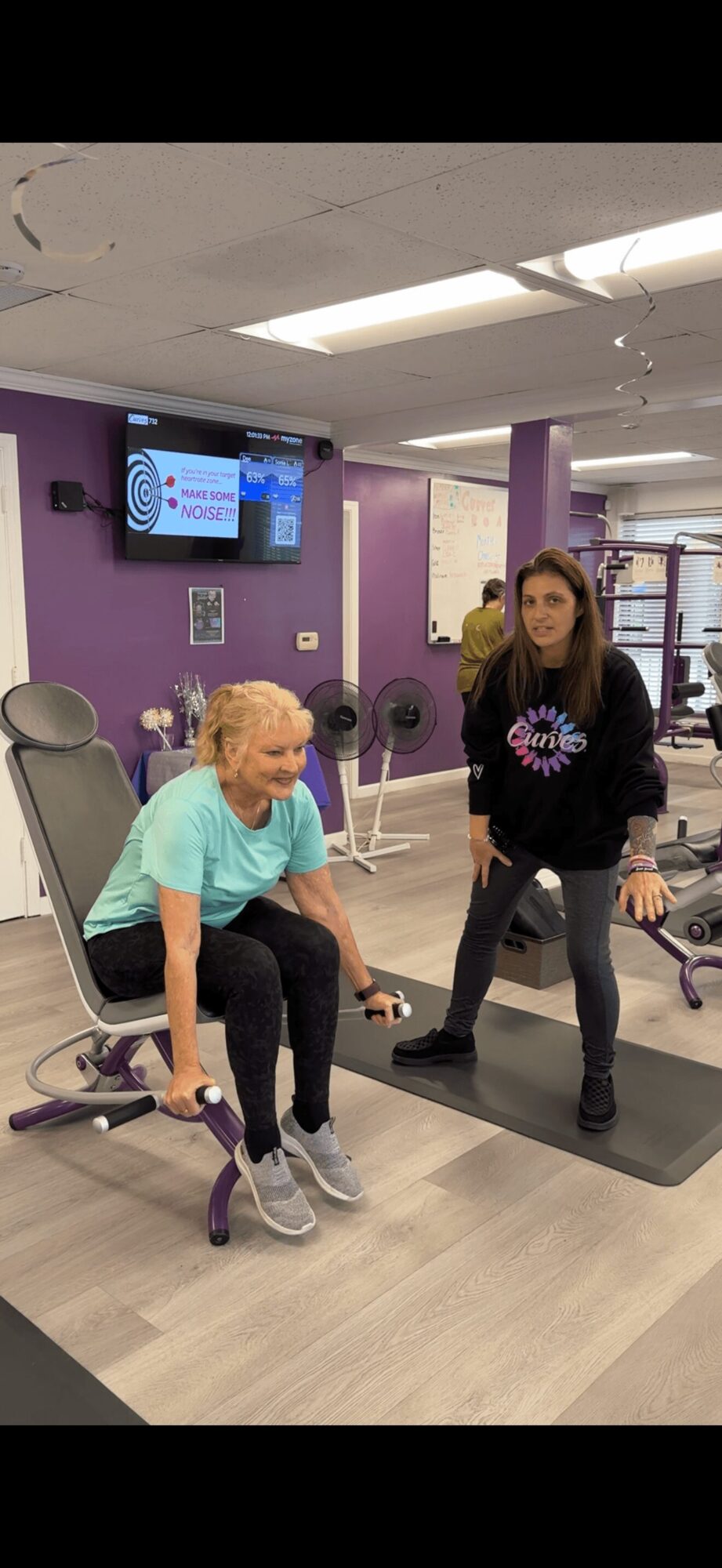Two women in a rehabilitation center, one sitting in a chair and the other standing nearby, with exercise equipment around.