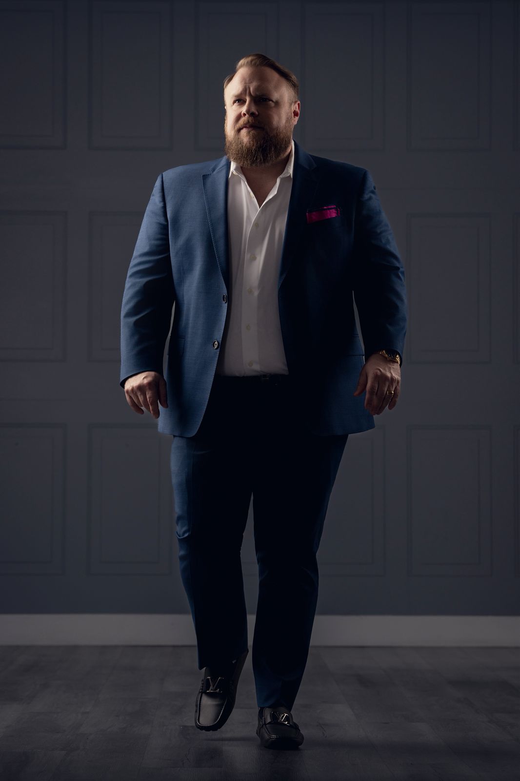Man in a blue suit walking towards the camera, dark background, serious expression, well-lit face and suit, full body