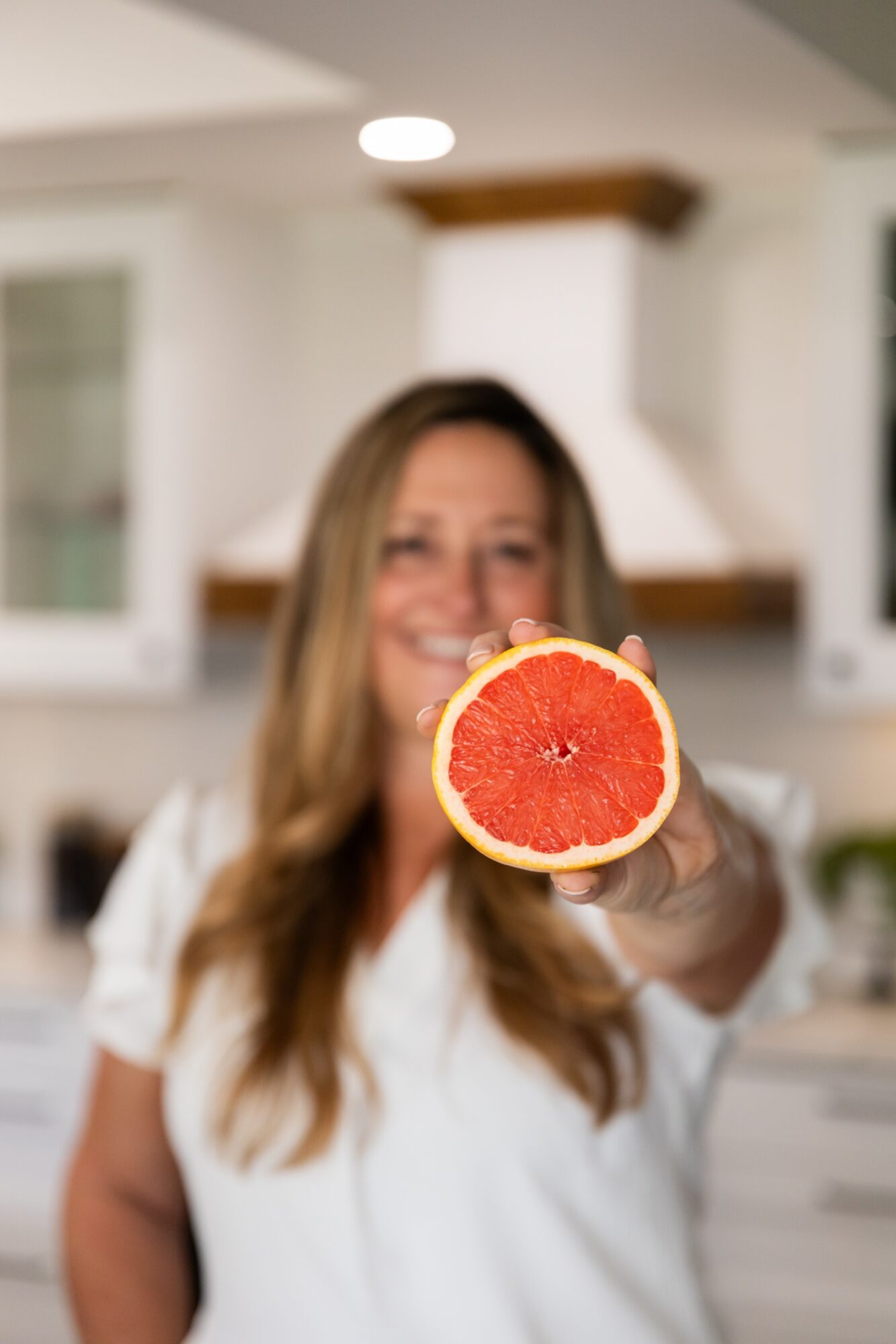 Woman holding a halved grapefruit towards the camera in a kitchen, smiling, with blurred background.
